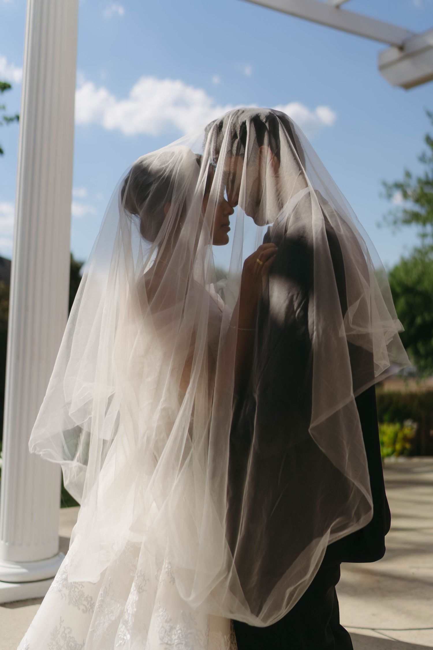 Bride and groom sharing intimate moment under flowing wedding veil outdoors at ceremony.