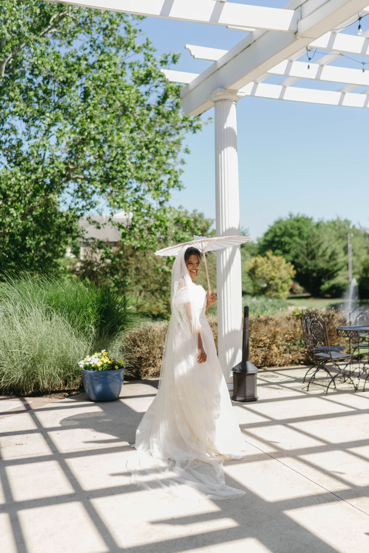 Bride in white gown and veil stands under pergola in sunlit outdoor garden venue on wedding day.