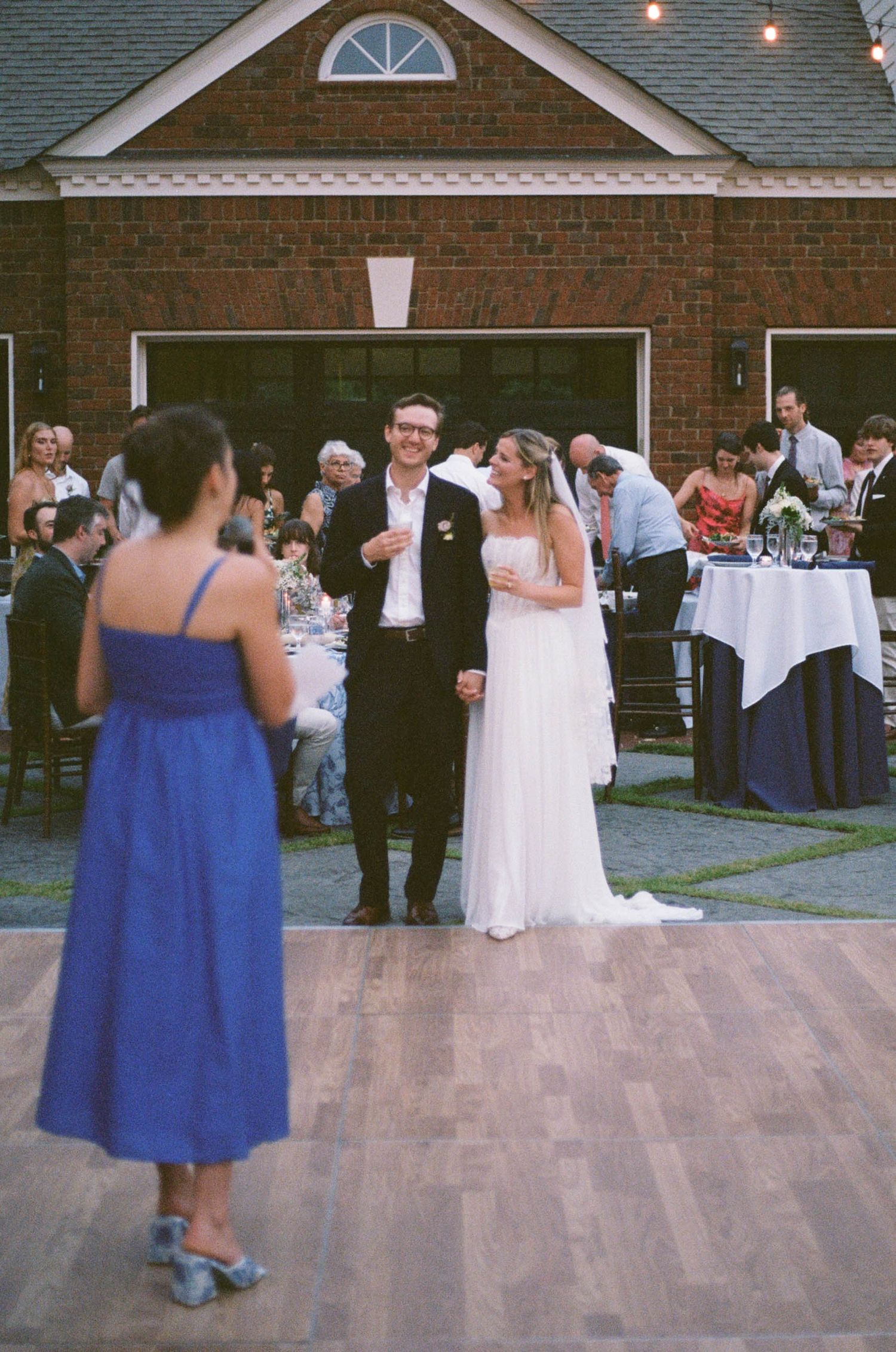 Bride and groom smile on outdoor dance floor at elegant brick venue wedding reception with guests seated nearby.