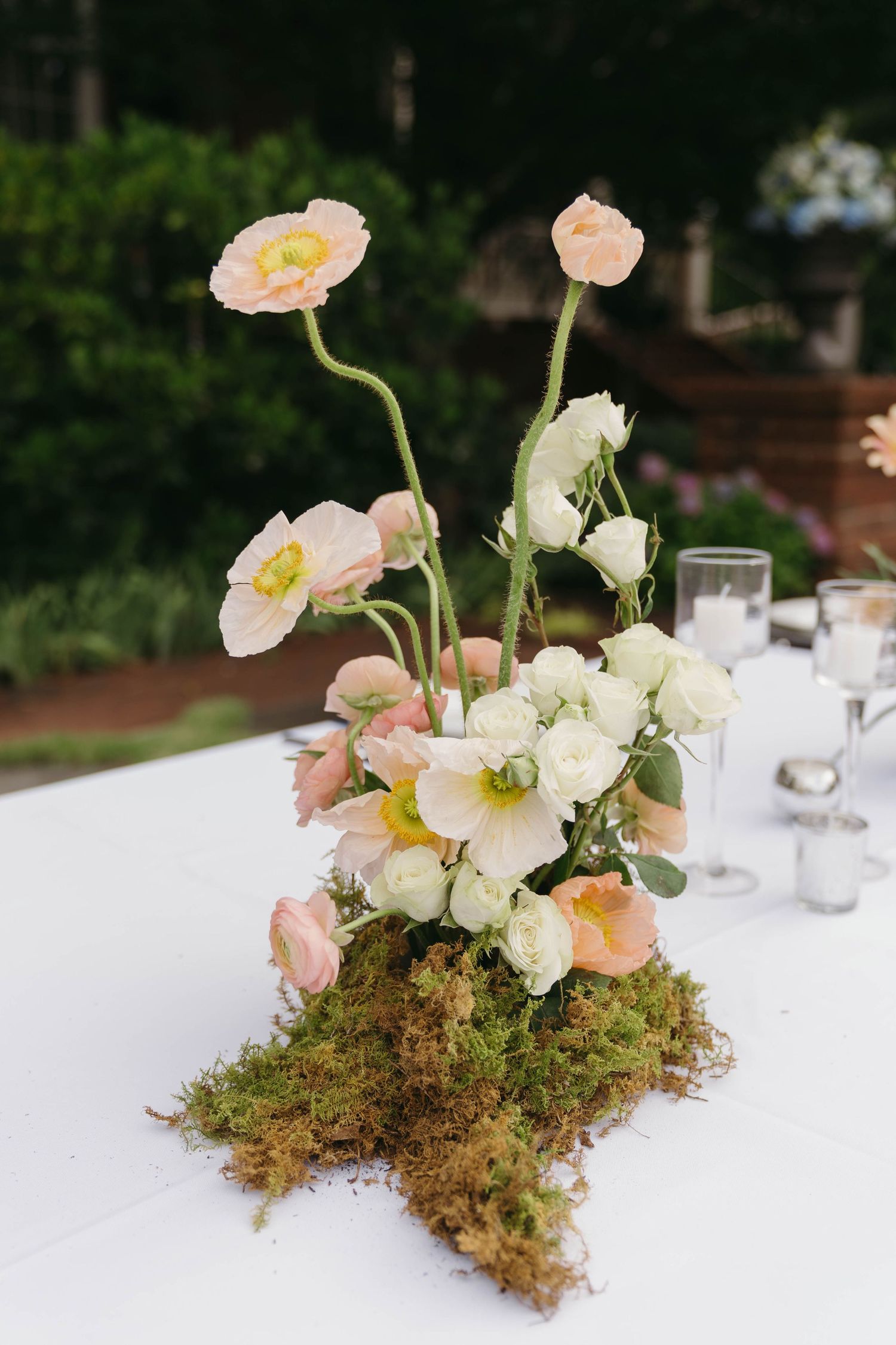 Elegant floral centerpiece with pink poppies and white roses arranged on a moss base for an outdoor event.