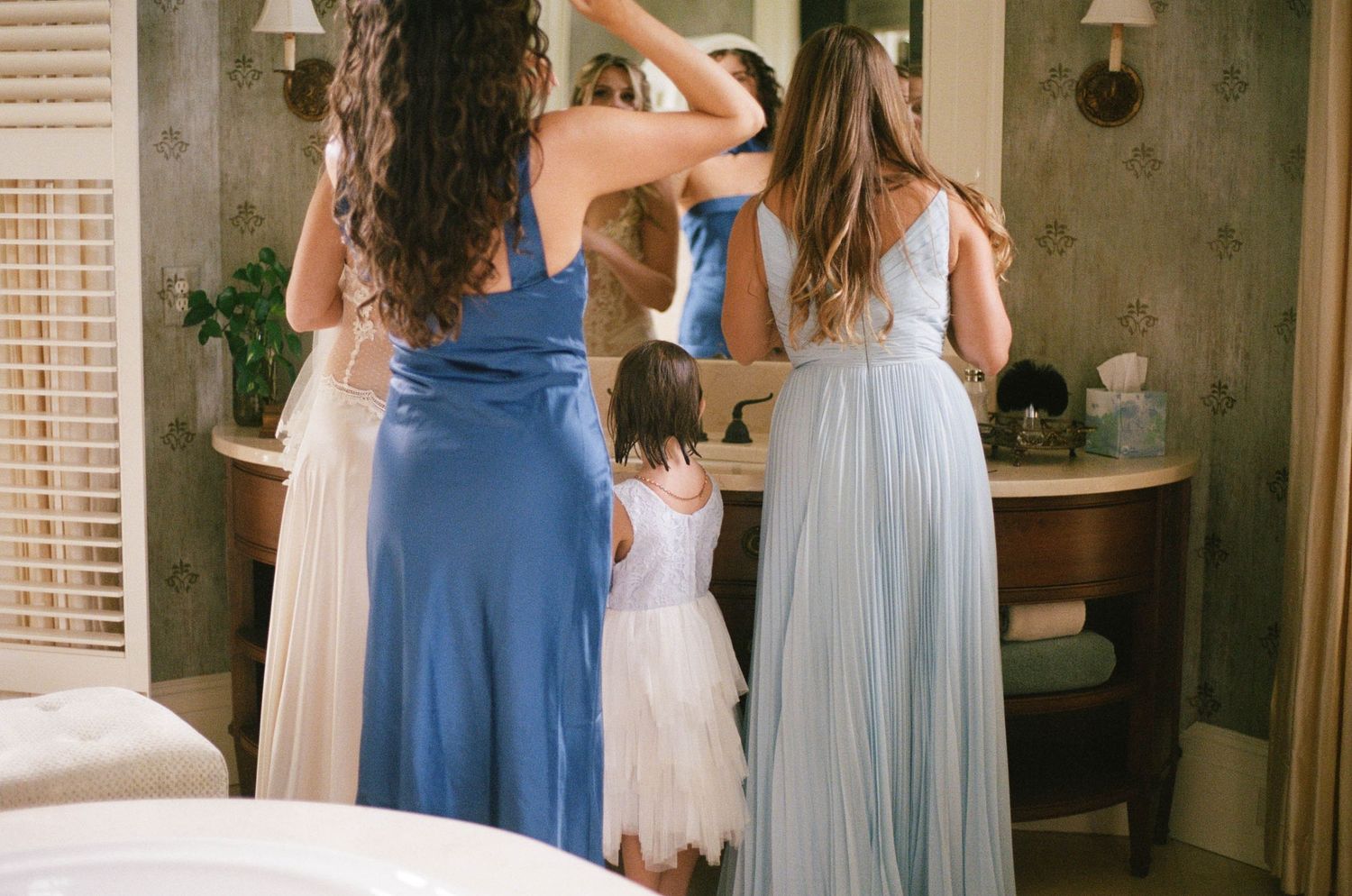 Bridesmaids in blue gowns and flower girl prepare together in an elegant dressing room mirror.