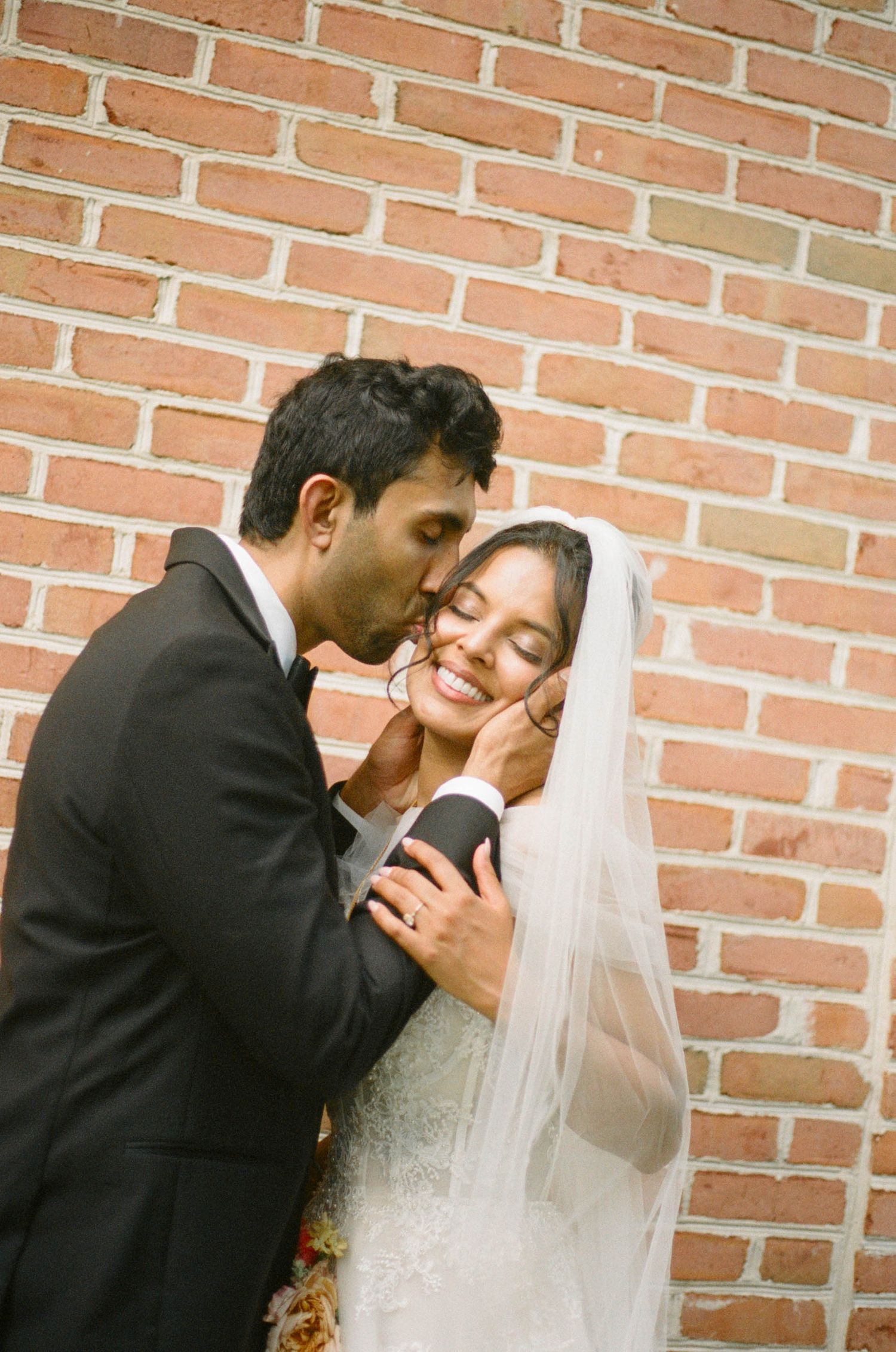 Groom tenderly kisses brides cheek as she smiles against a red brick wall on their wedding day.