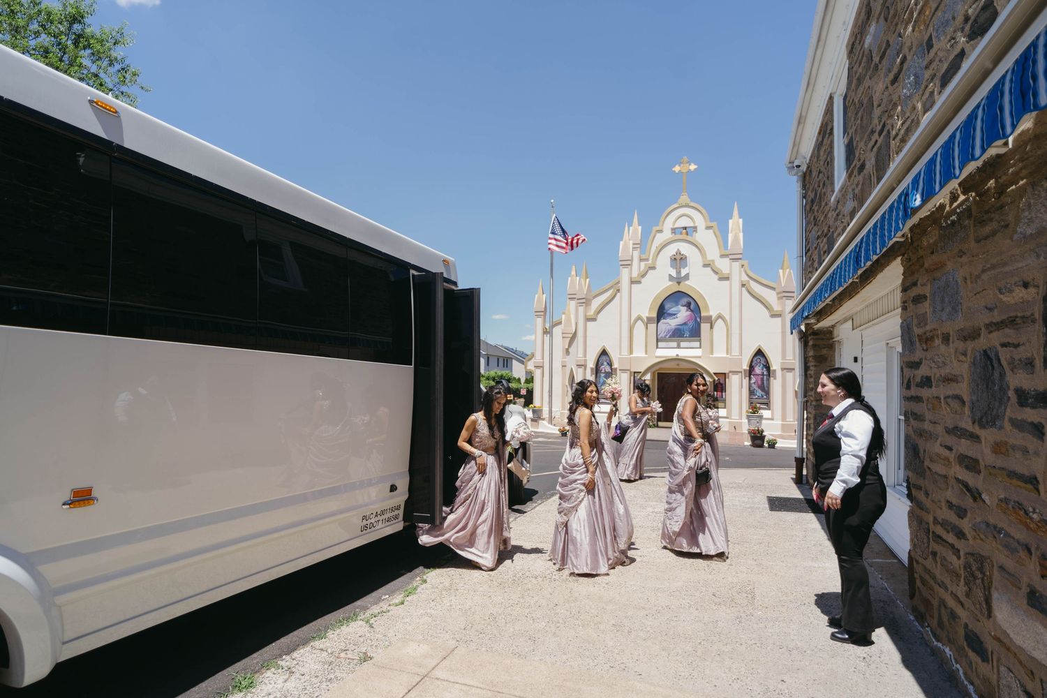 Bridesmaids in lavender sarees boarding party bus outside ornate Catholic church on sunny wedding day.