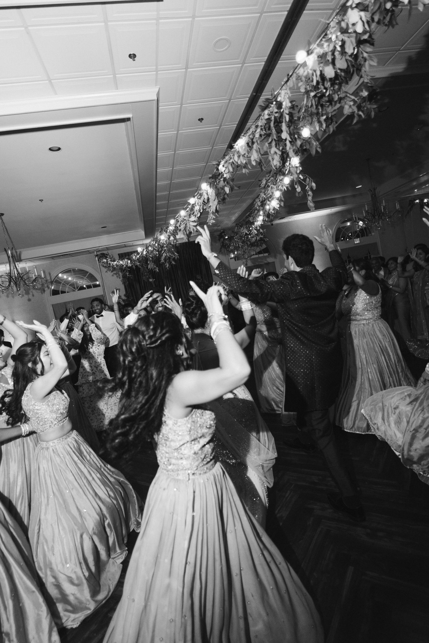 Black and white photo of wedding guests in formal gowns dancing joyfully at a reception under floral decorations.