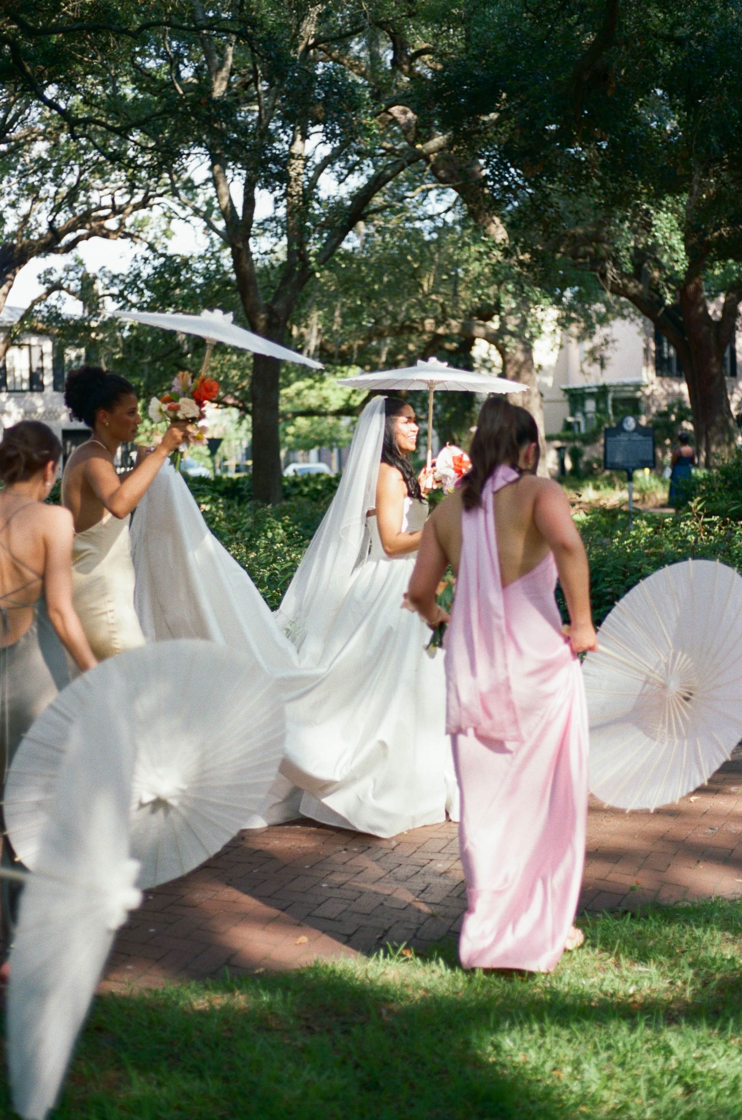 Wedding party members pose with white parasols on a brick pathway.