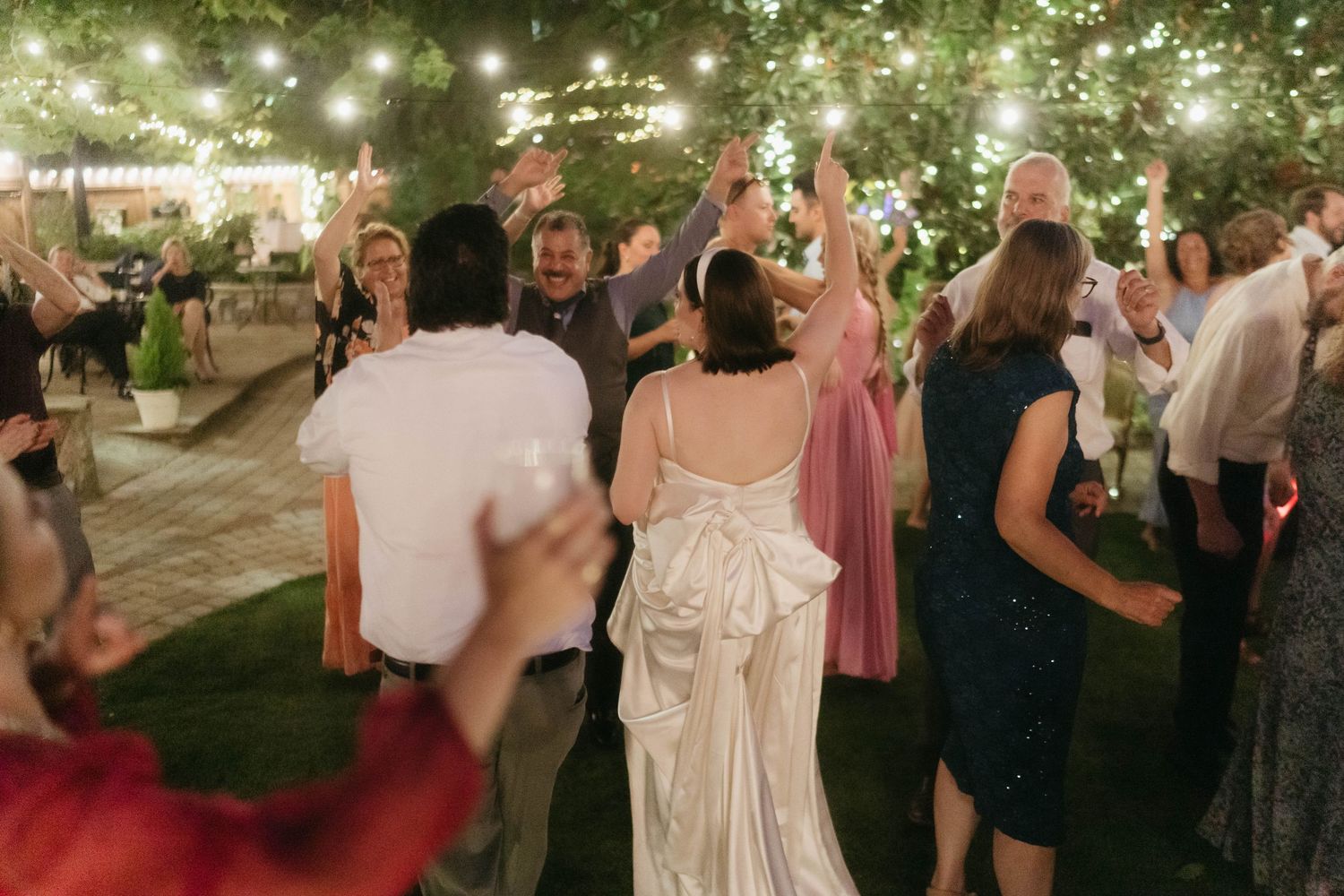 Wedding guests dance together under twinkling string lights at an outdoor evening reception.