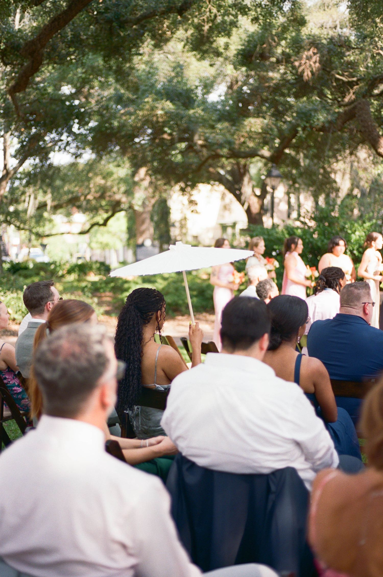 Wedding guests watch ceremony under oak trees while someone holds a white parasol.