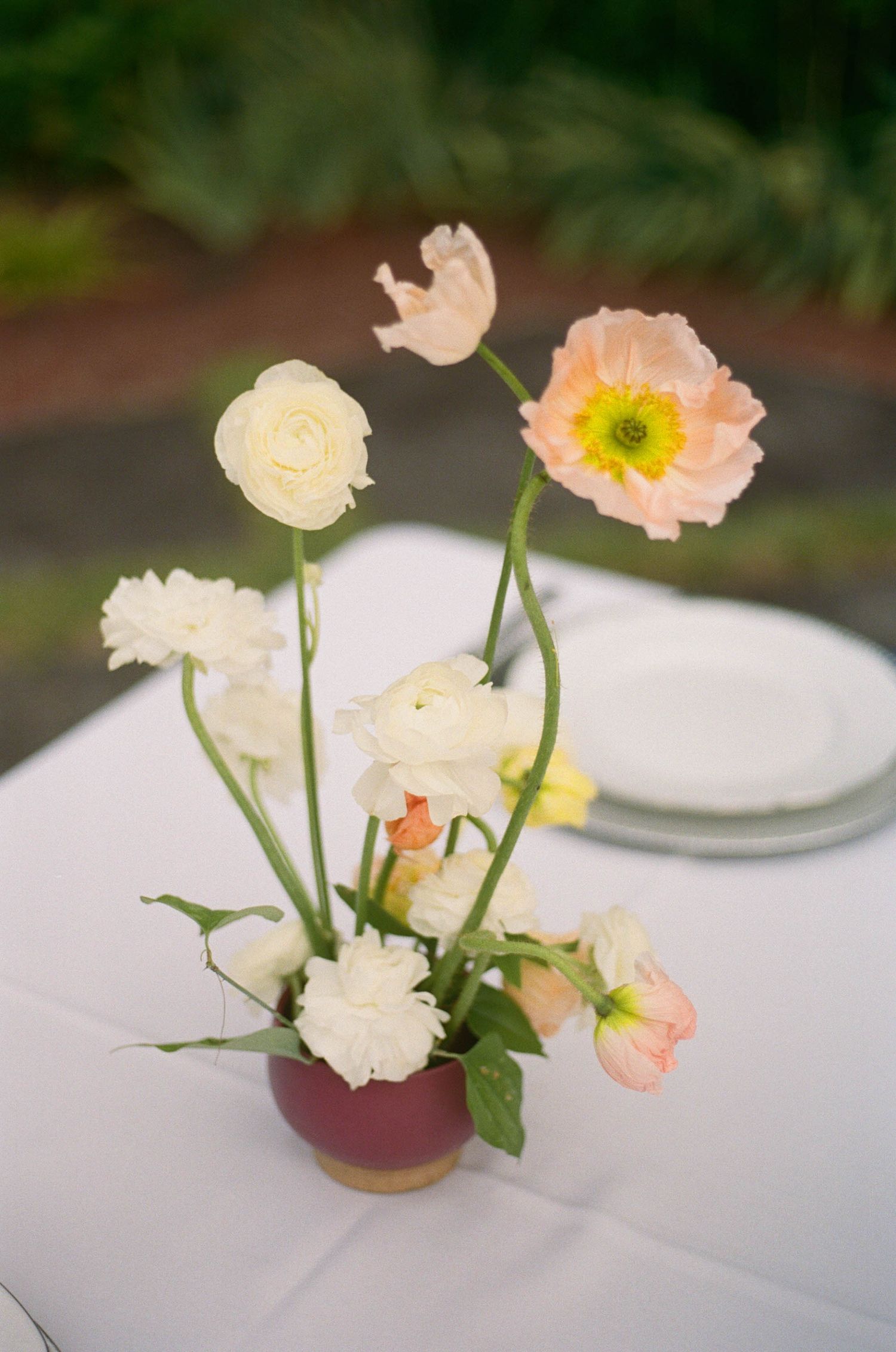 Small burgundy vase with white ranunculus and peach poppy flowers on a white tablecloth at an outdoor wedding reception.