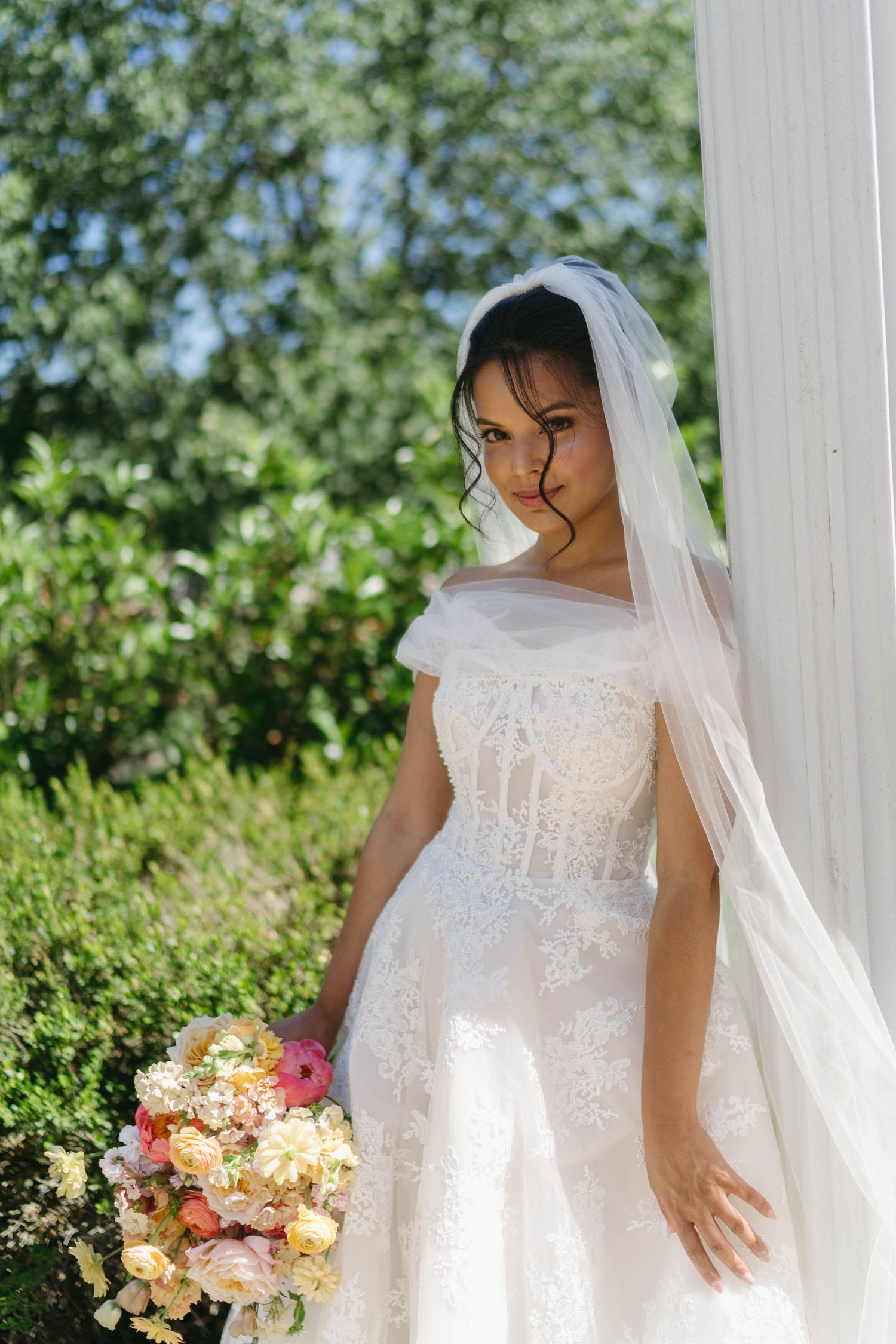 Elegant bride in lace wedding gown and veil holds colorful bouquet beside white column outdoors.