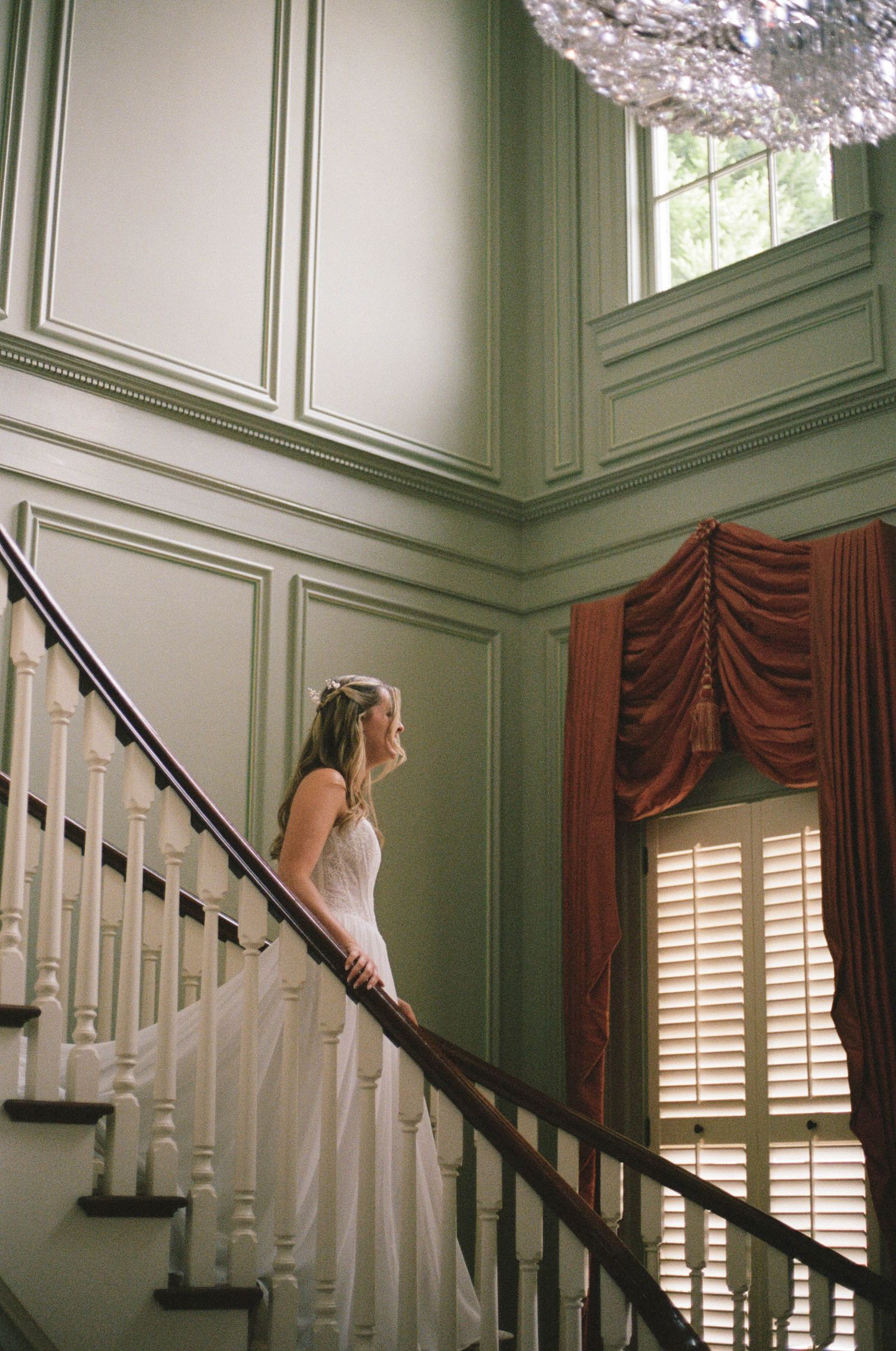 Bride in white gown stands on elegant staircase with green walls and red drapes.