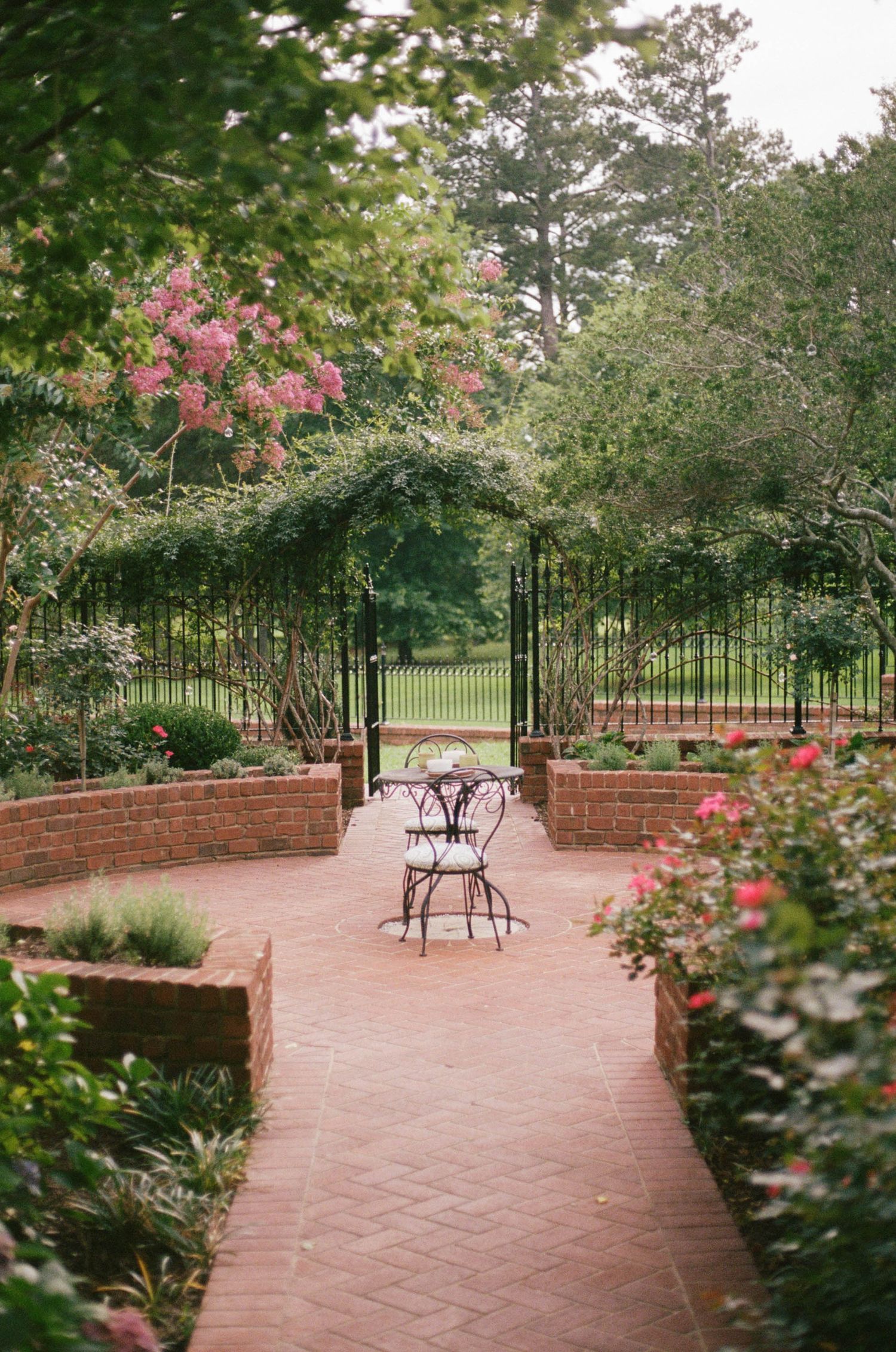 Brick garden path leads to ornate iron gate arbor with bistro table, surrounded by lush greenery and blooming pink flowers.