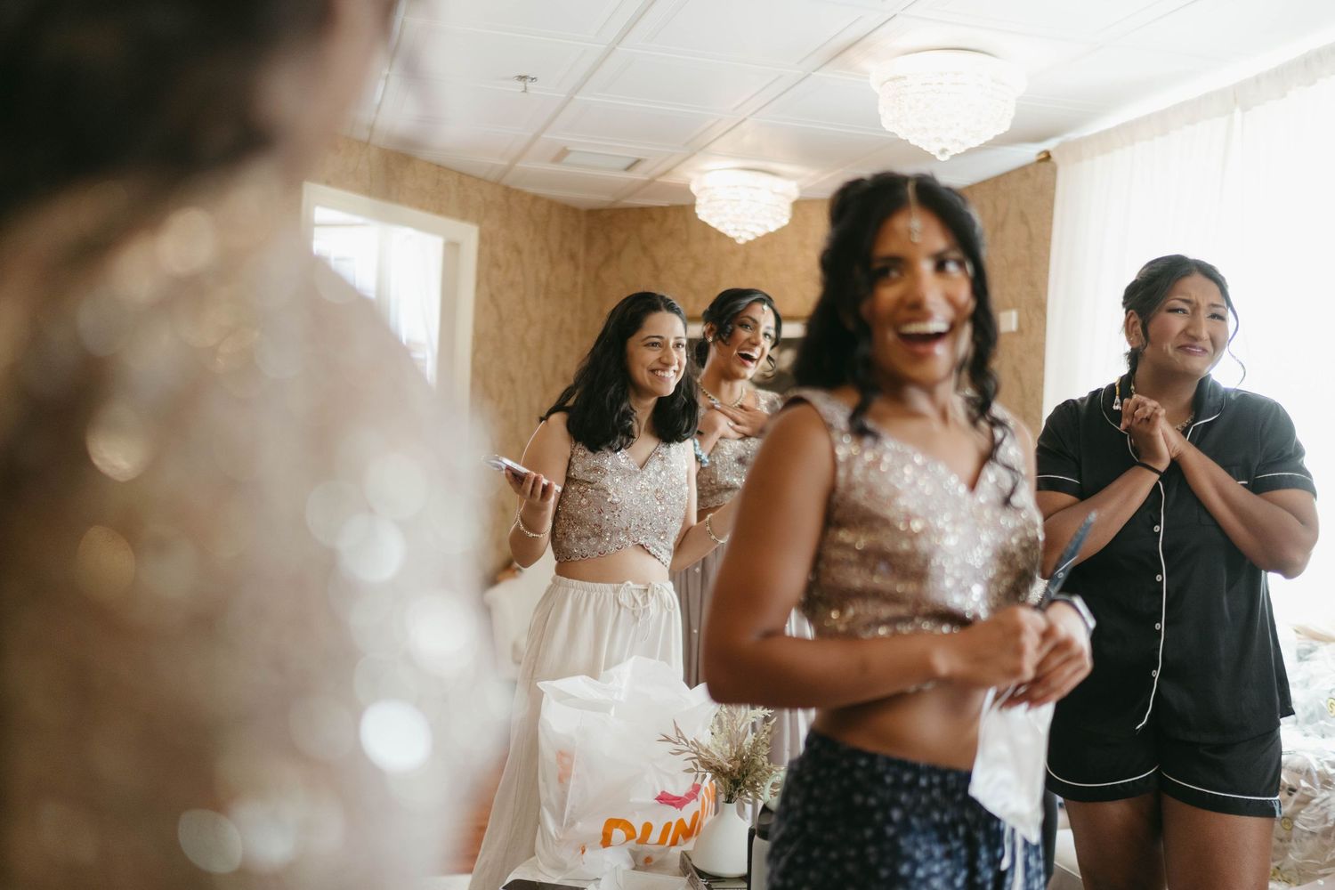 Bridesmaids react with joy and excitement as bride reveals her wedding dress during bridal preparations.