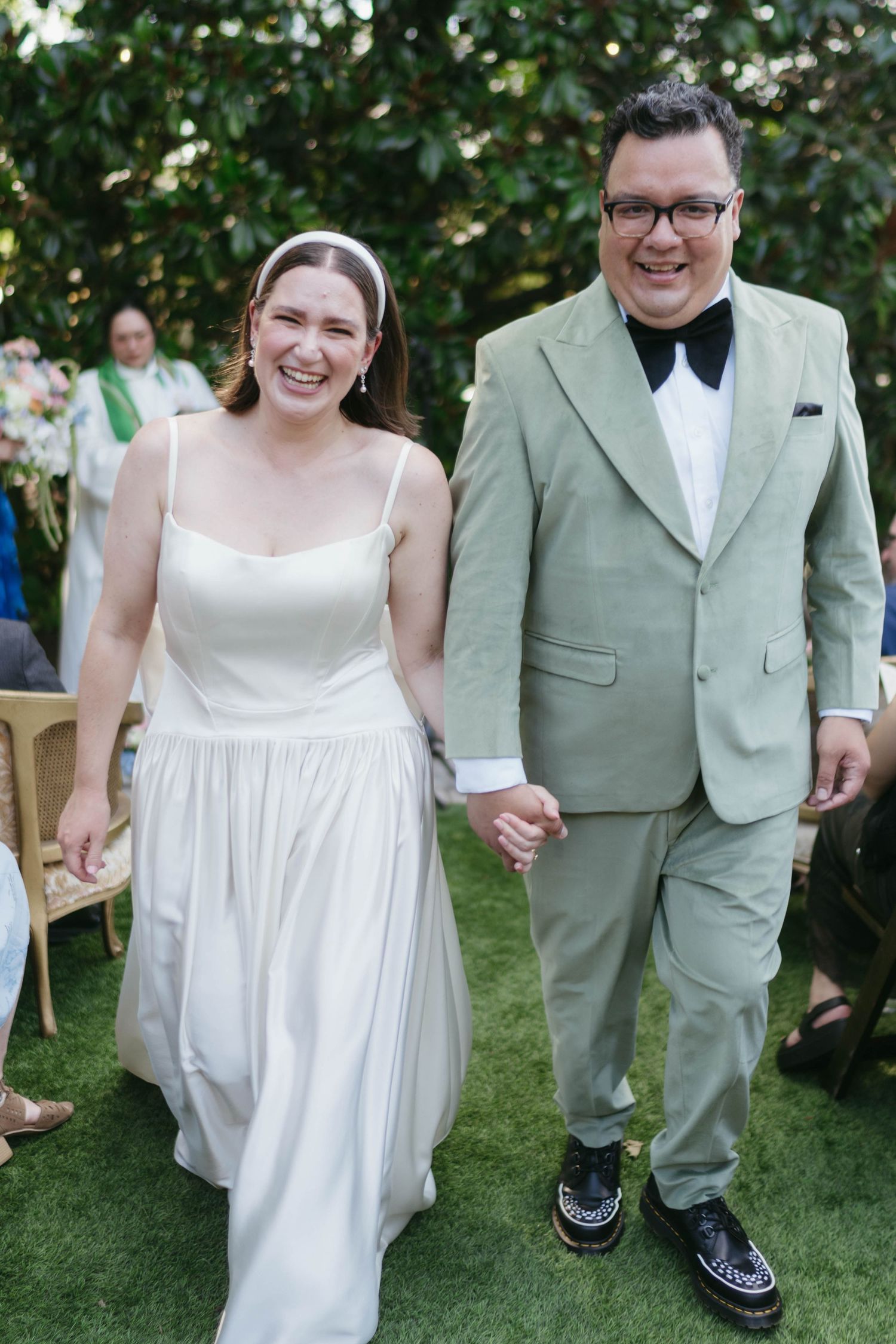 Wedding couple walks hand in hand across a garden lawn, with the bride in white and groom in sage green.