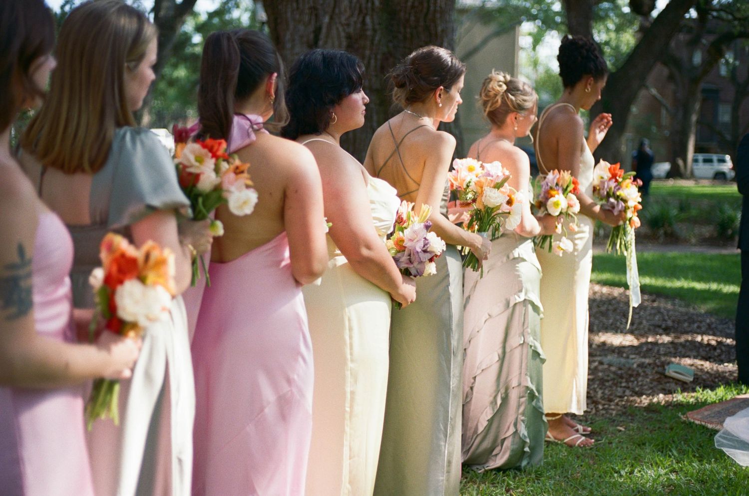 A row of bridesmaids in pastel dresses holding floral bouquets at an outdoor wedding.