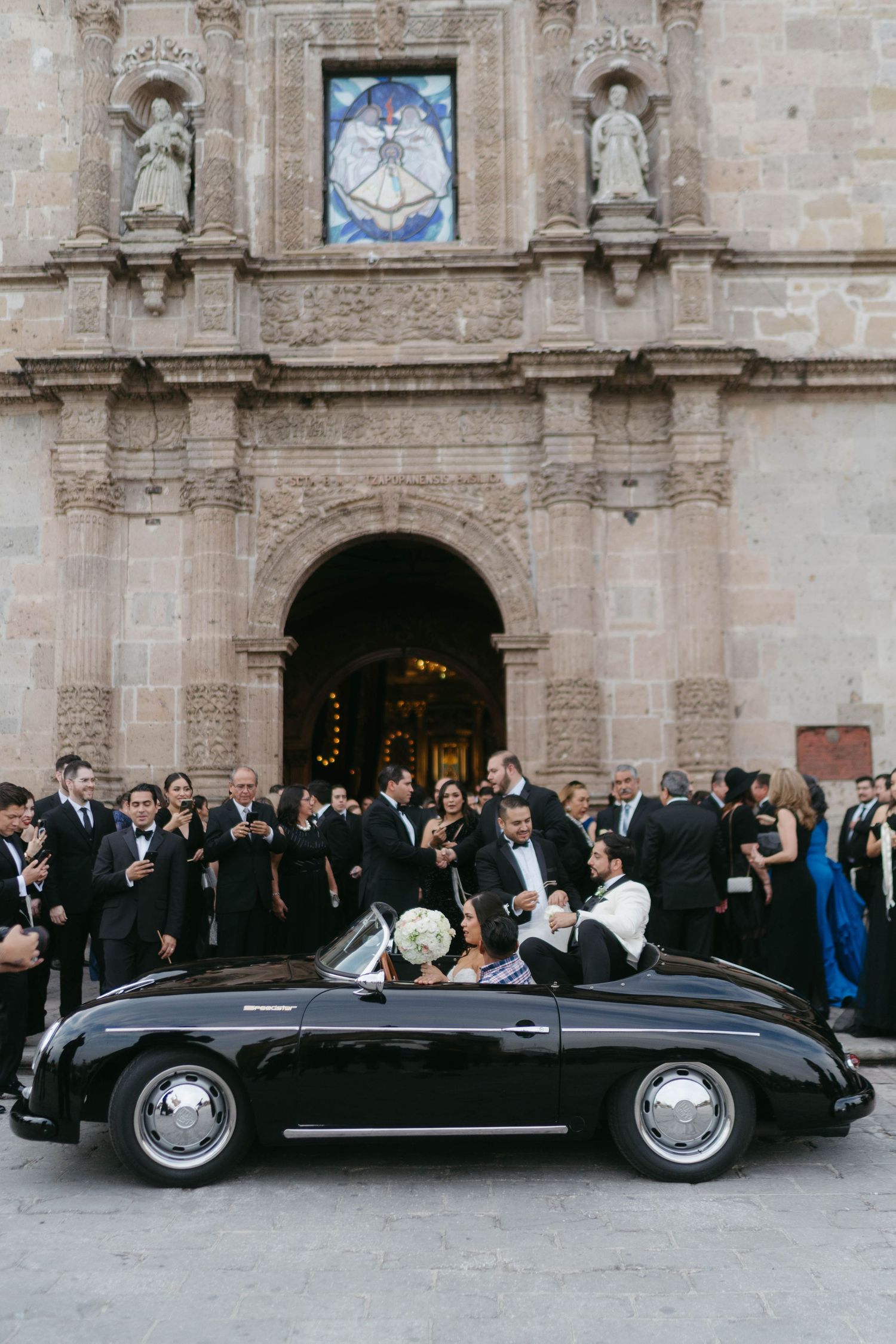 Classic black convertible car parked outside historic stone church with wedding guests.
