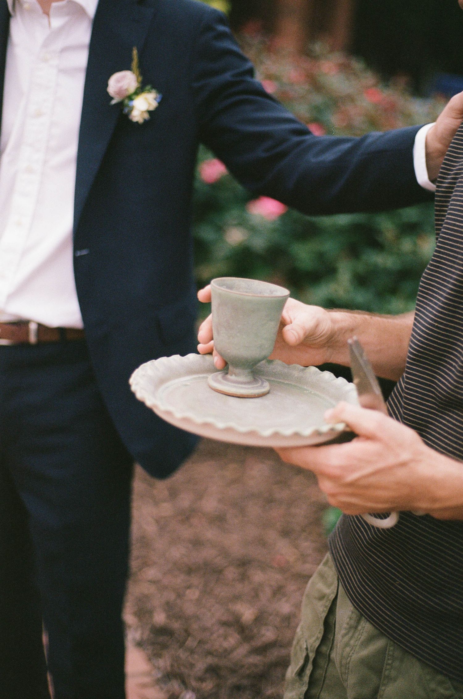 Groom in navy suit with boutonniere receives ceramic cup on rustic plate during outdoor wedding ceremony.