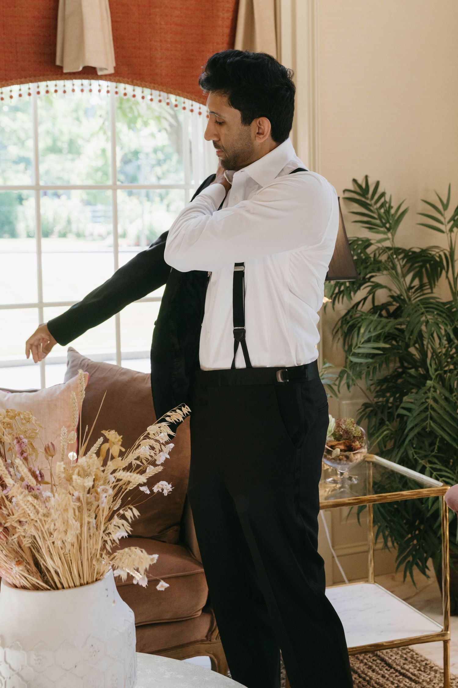 Groom in white shirt and suspenders putting on his tuxedo jacket while getting ready for his wedding day.