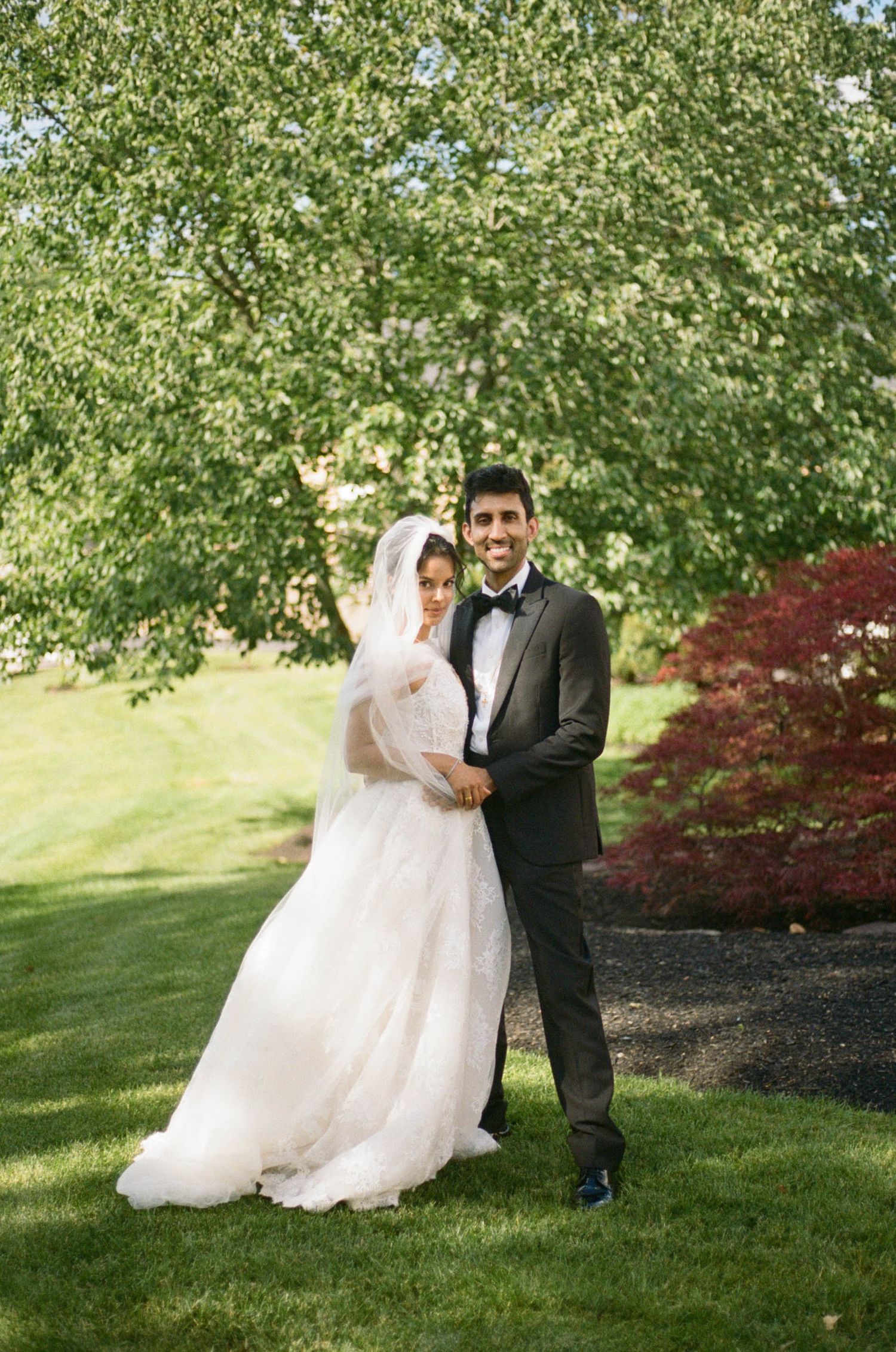 Bride and groom pose together in a lush green garden on their wedding day, smiling warmly.