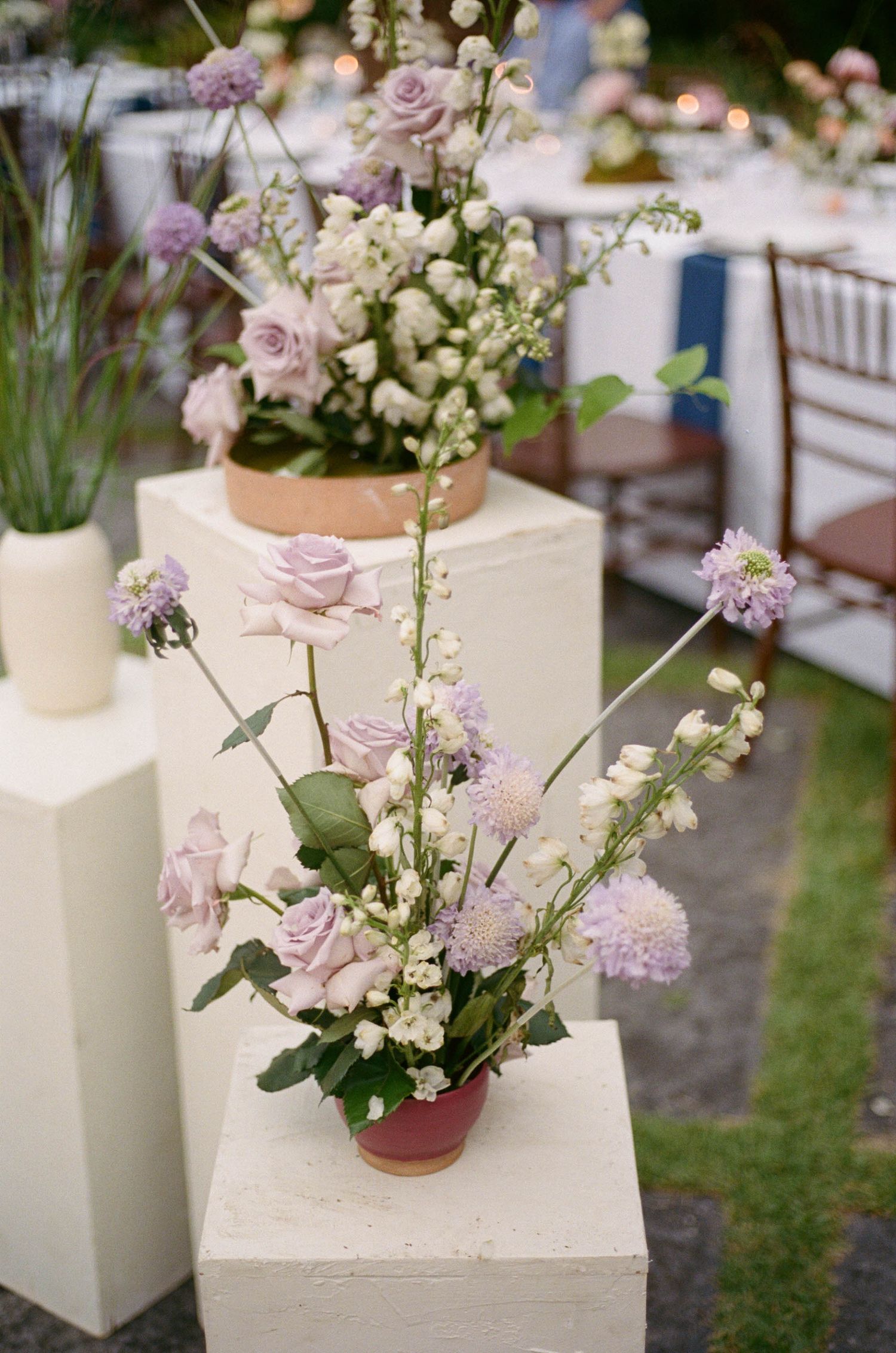 Elegant lavender and white floral arrangements in pink pots displayed on white pedestals at a wedding reception.