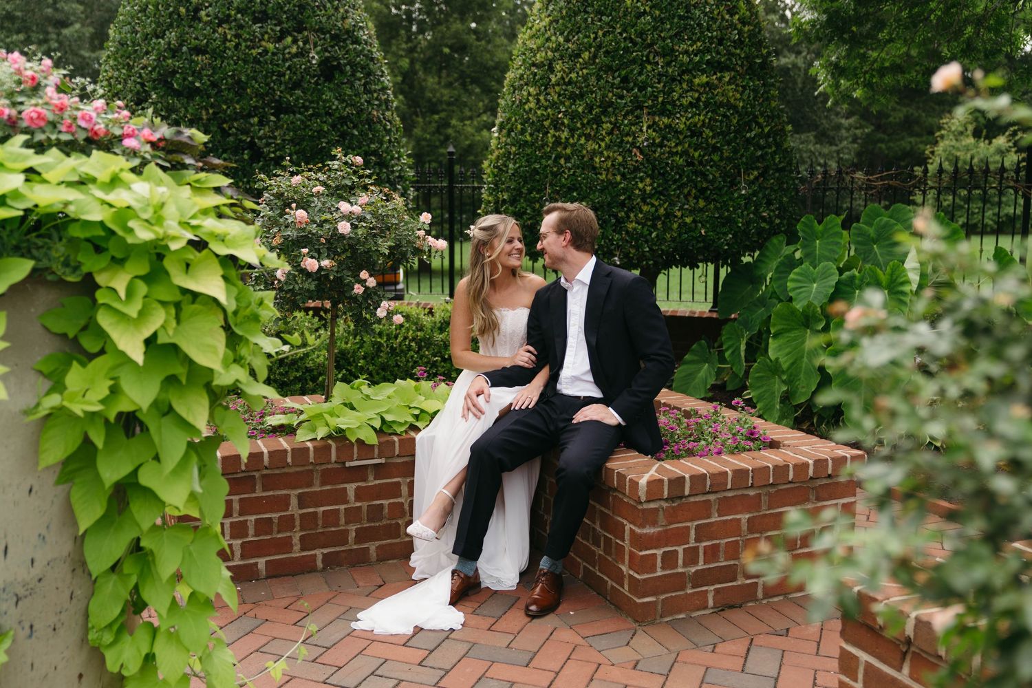 Bride and groom sit together on a brick garden bench surrounded by lush greenery and roses.
