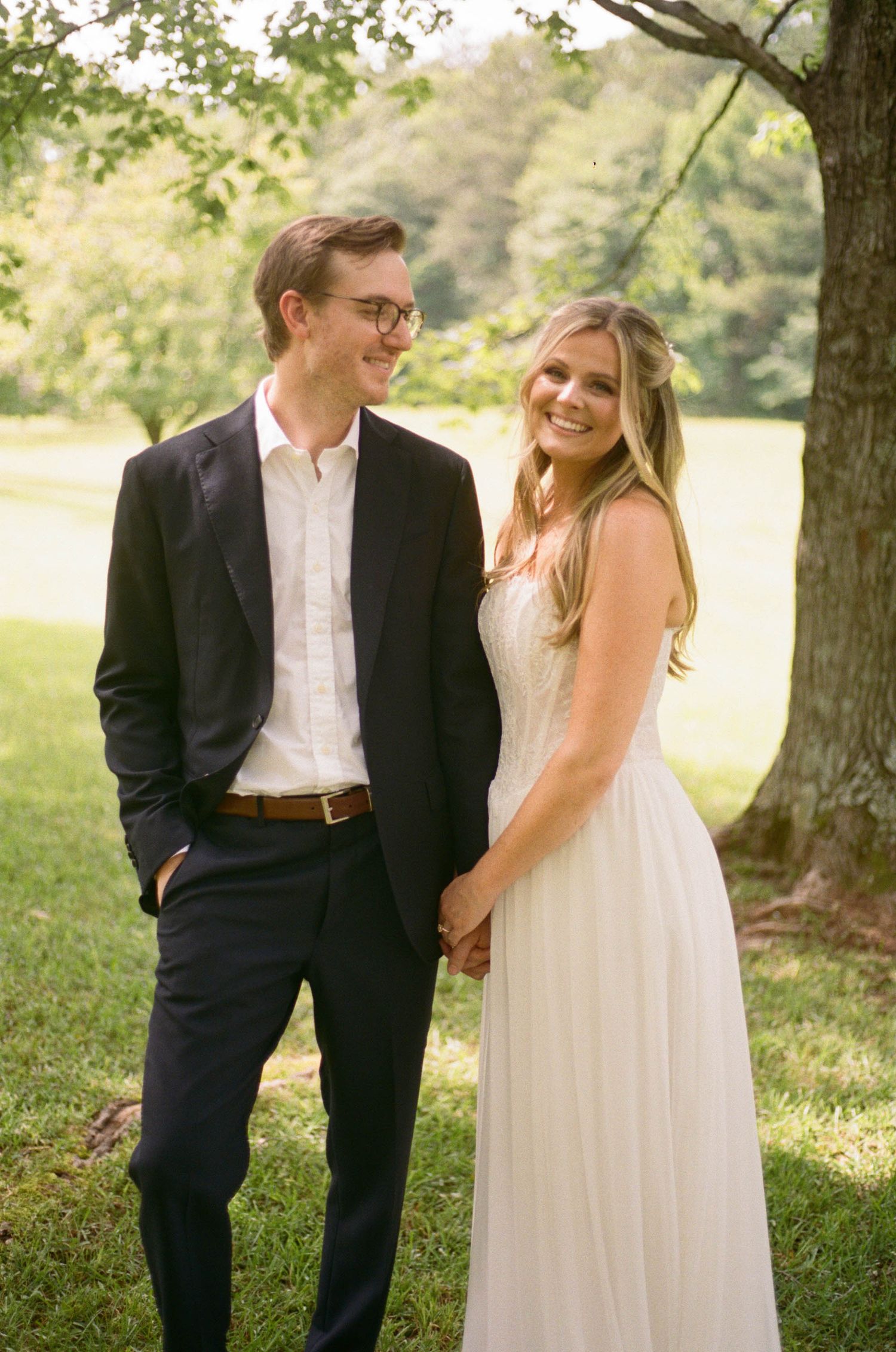Happy bride and groom holding hands outdoors under a tree, bride in flowing white dress, groom in black suit.