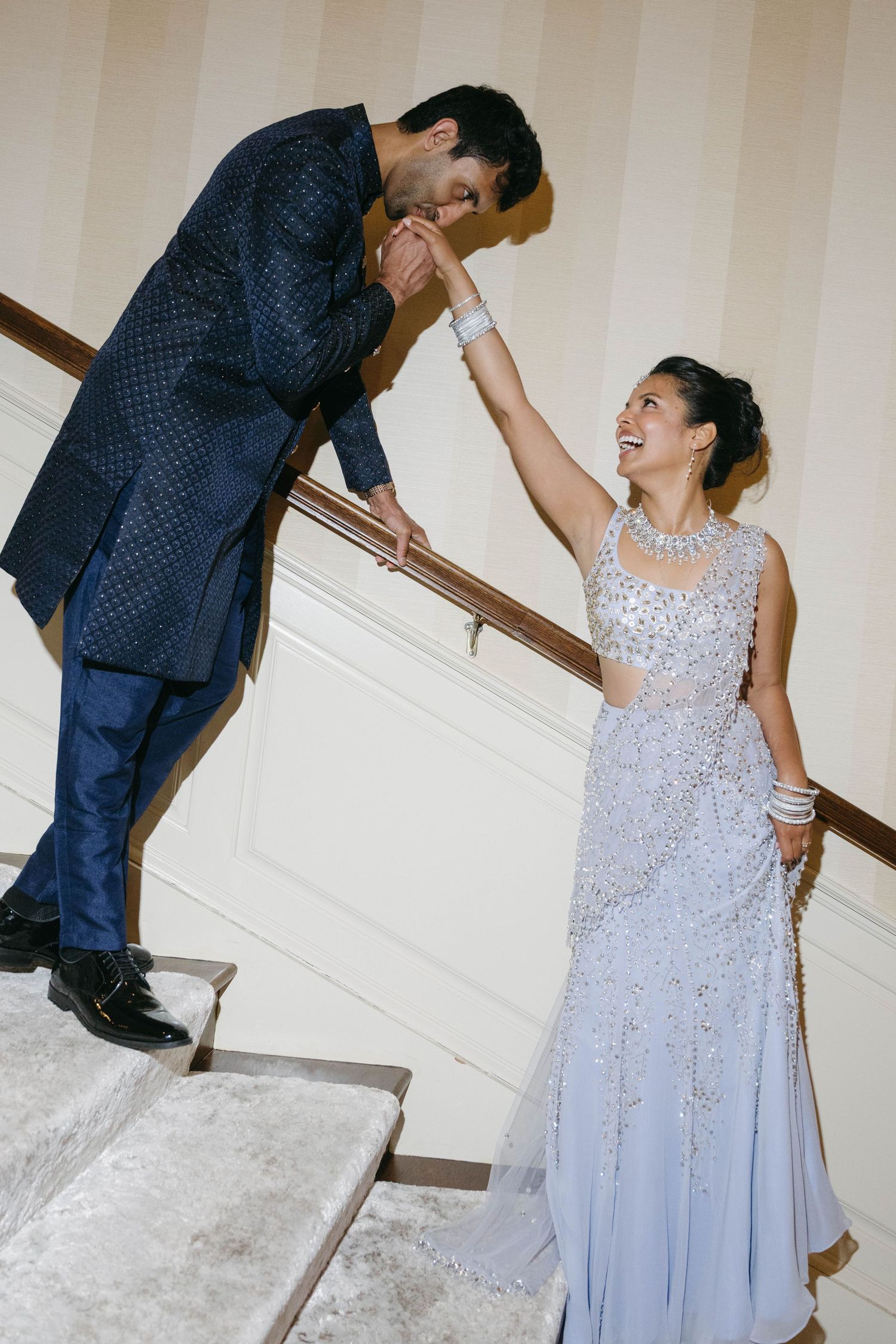 A groom in blue Indian attire kisses the hand of his bride in a silver embellished lehenga on stairs.