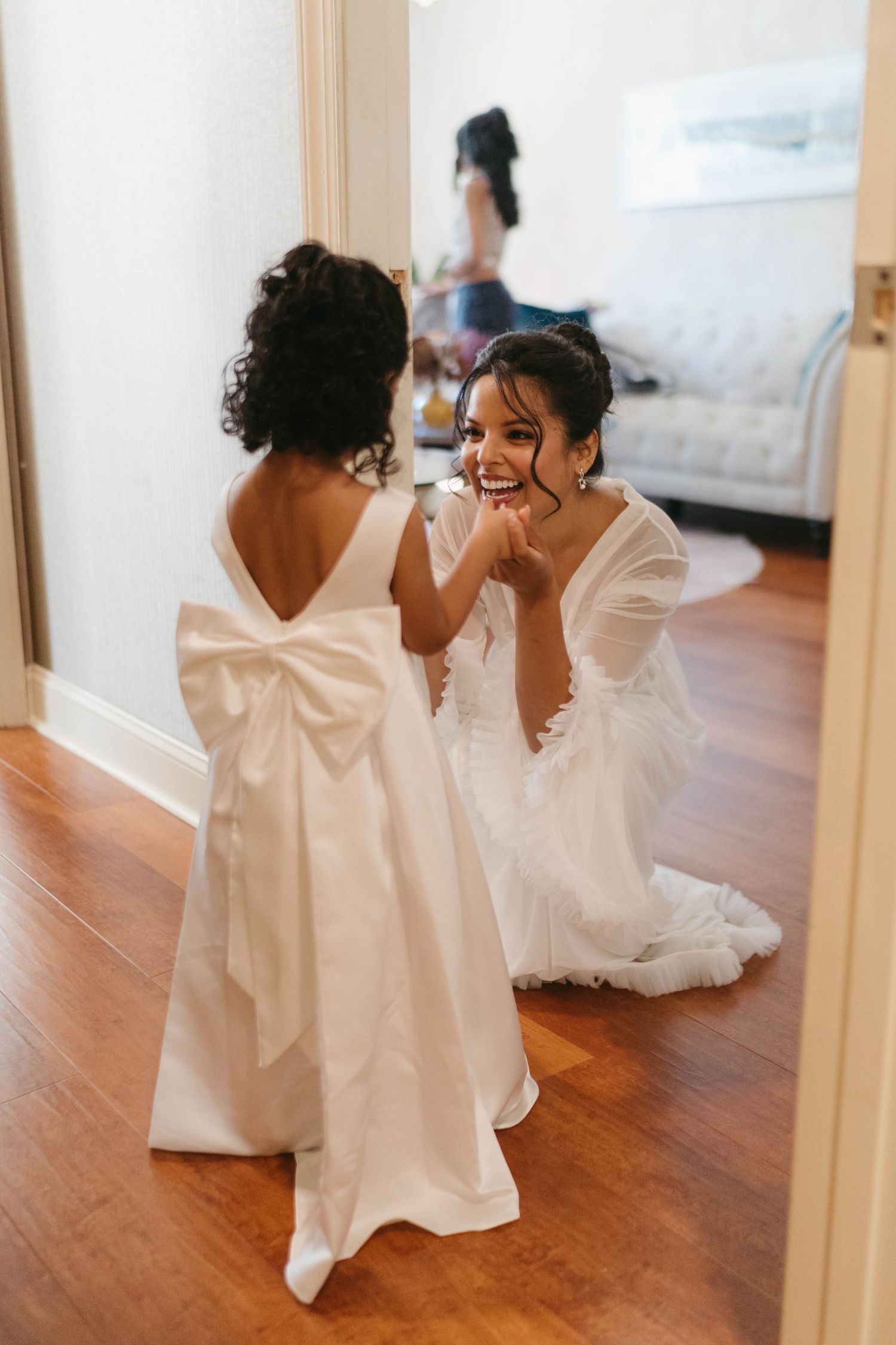 Smiling bride in white gown kneels to interact with flower girl wearing white dress with bow.