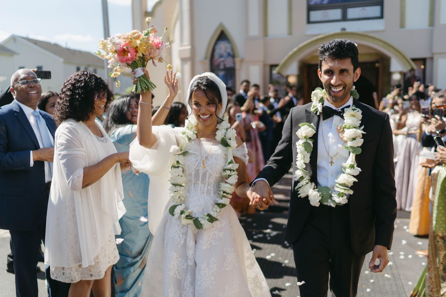Happy bride and groom wearing floral leis walk out of church as guests celebrate around them.