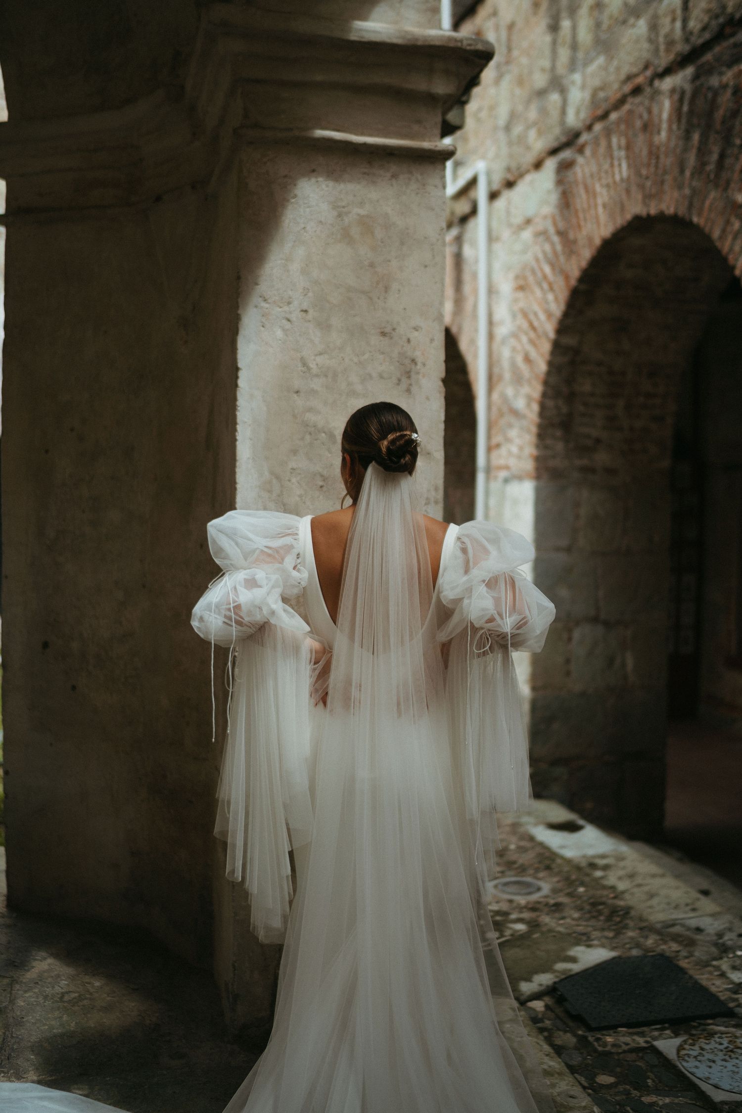 A dramatic backlit wedding dress with puff sleeves and long veil in a stone corridor.