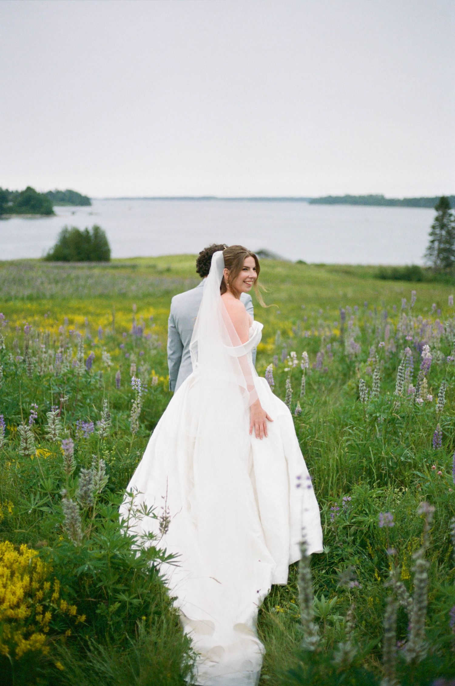 Bride in flowing white dress among purple lupines with coastal scenery in the background.