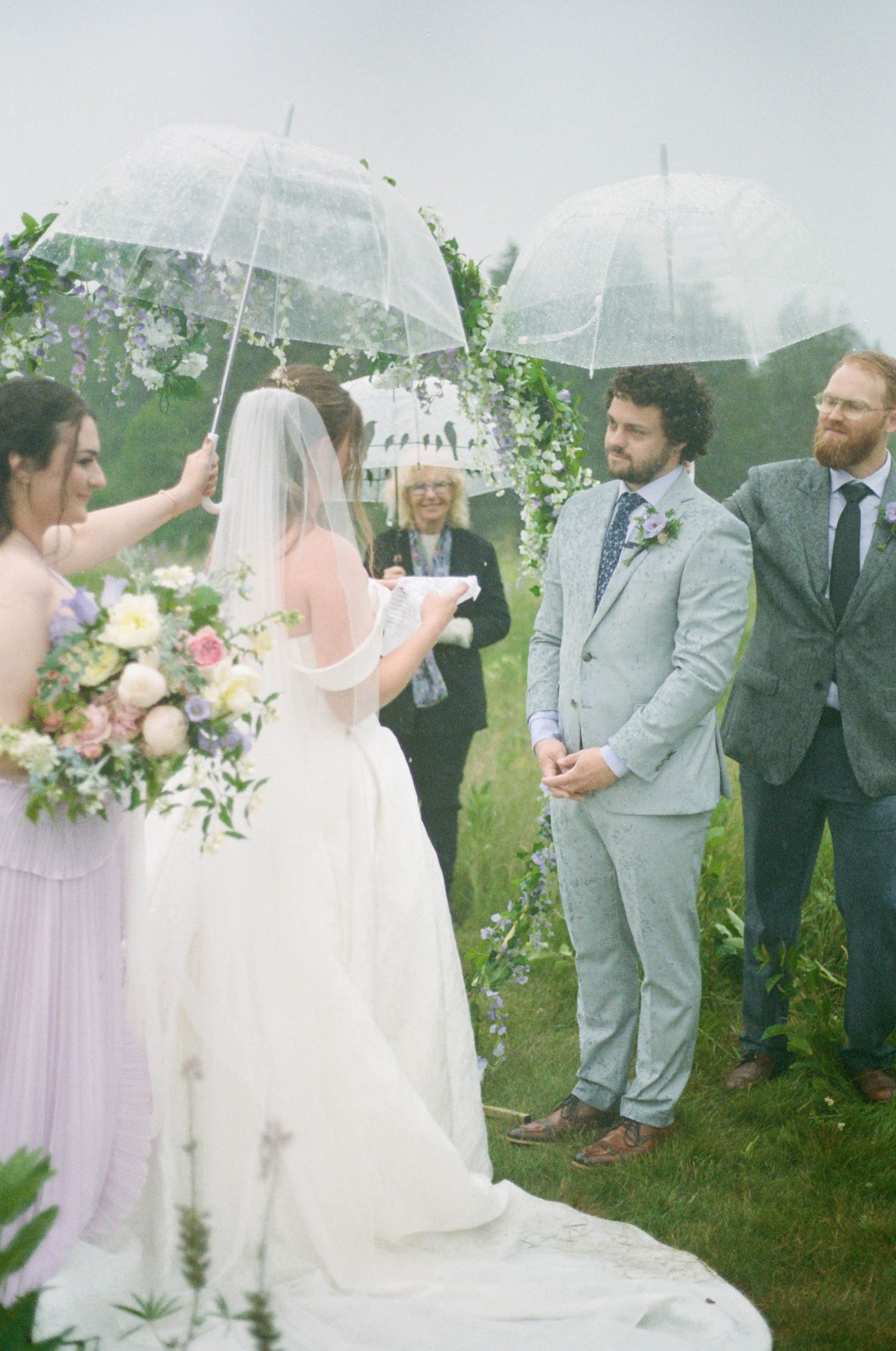 Outdoor wedding ceremony with clear umbrellas and floral arch in wildflower field.