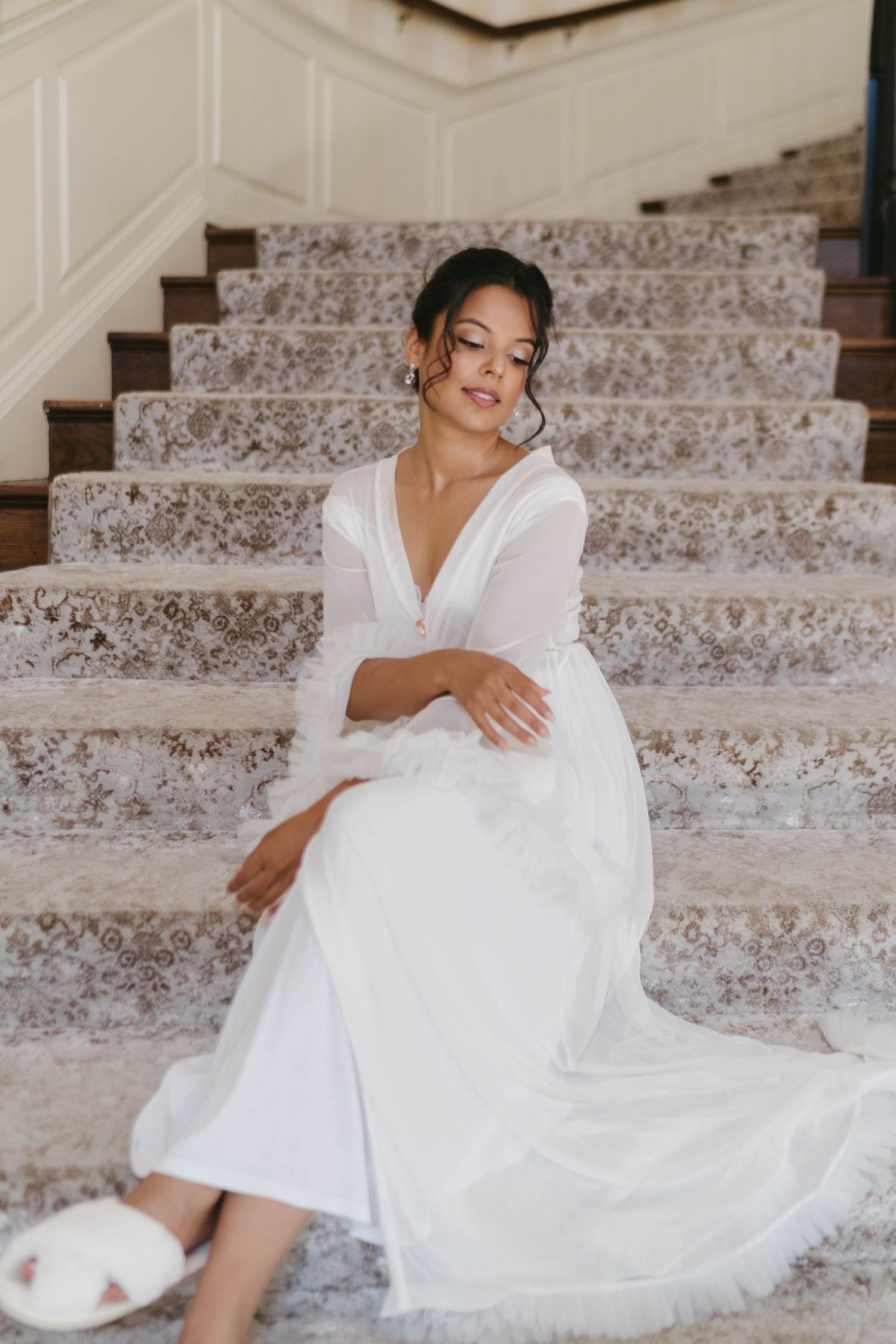 Bride in white long-sleeve gown sitting gracefully on marble staircase with lace carpet runner.