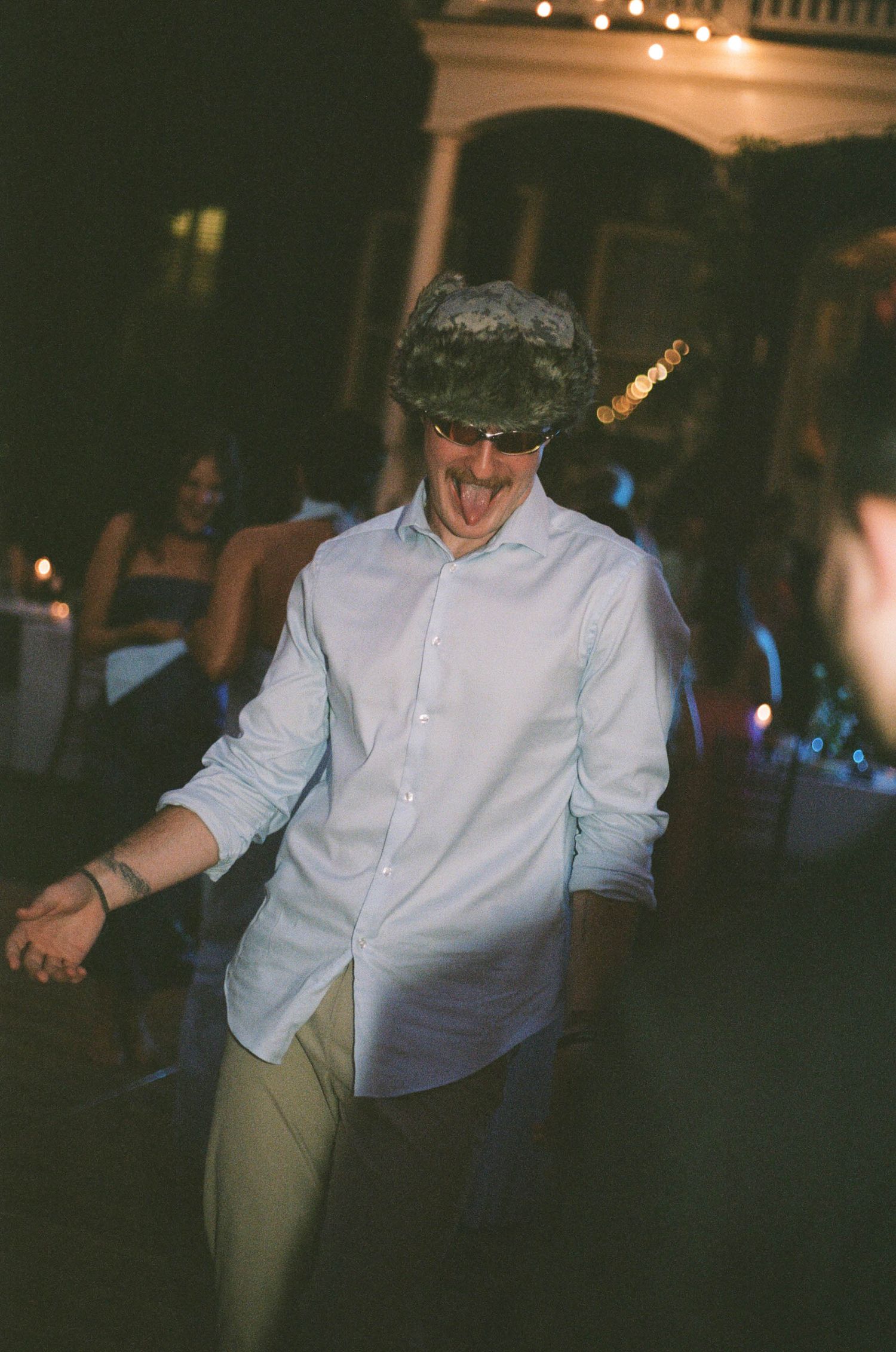 Wedding guest dances energetically wearing a hat and sunglasses at a lively evening reception.
