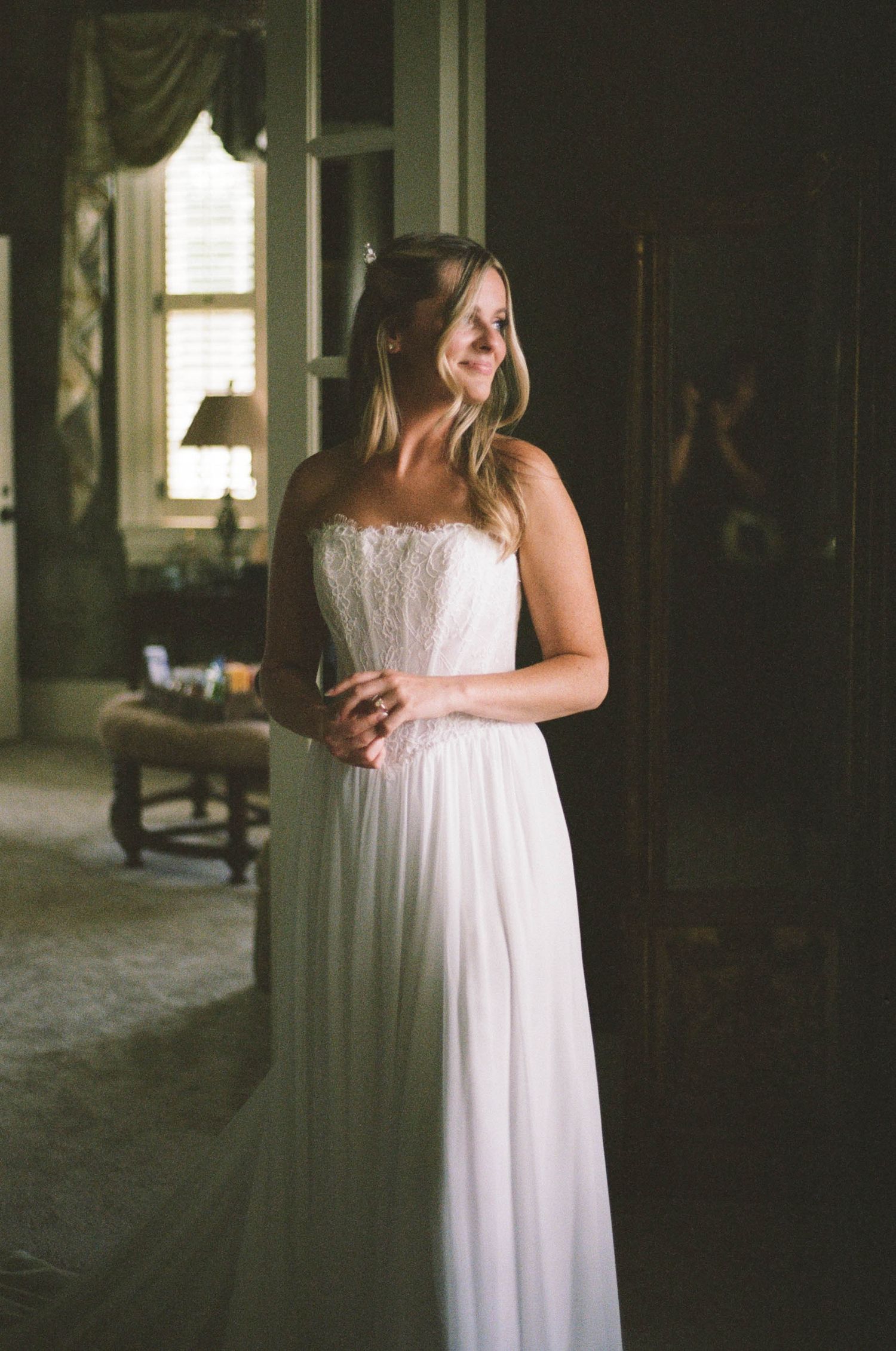 Bride smiling in elegant strapless white lace and chiffon wedding gown standing in a softly lit room.