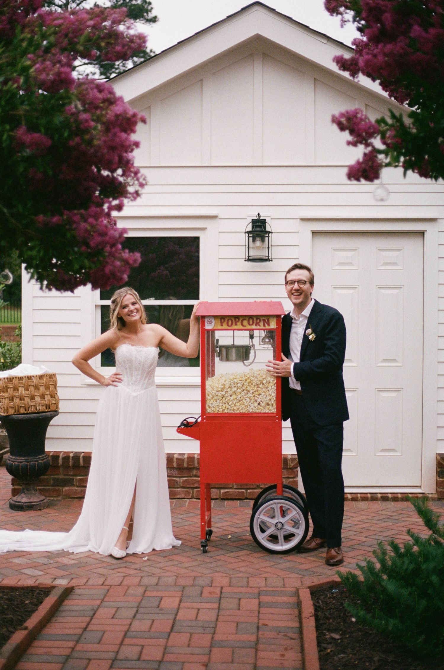 Bride and groom smiling beside a red popcorn cart outside a white barn venue with blooming pink flowers.