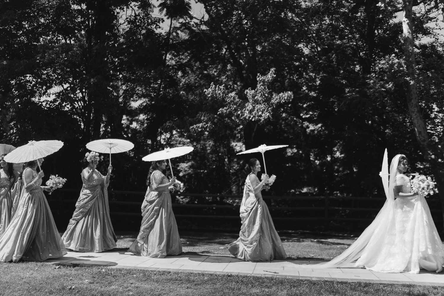 Black and white photo of bride and bridesmaids walking outdoors carrying white parasol umbrellas.