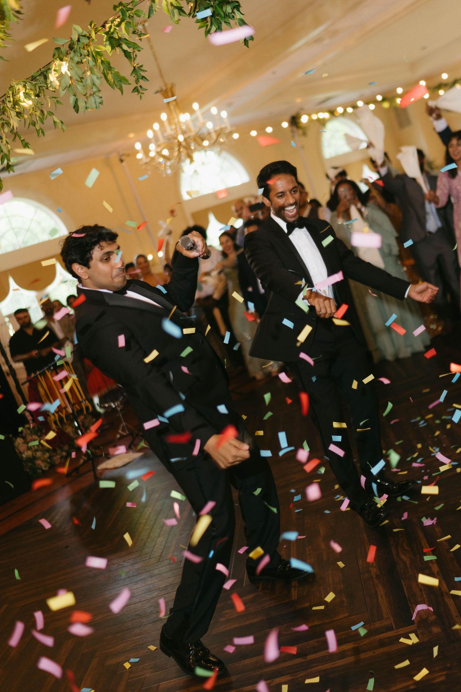 Two groomsmen in tuxedos dance joyfully on wedding reception dance floor surrounded by colorful confetti.