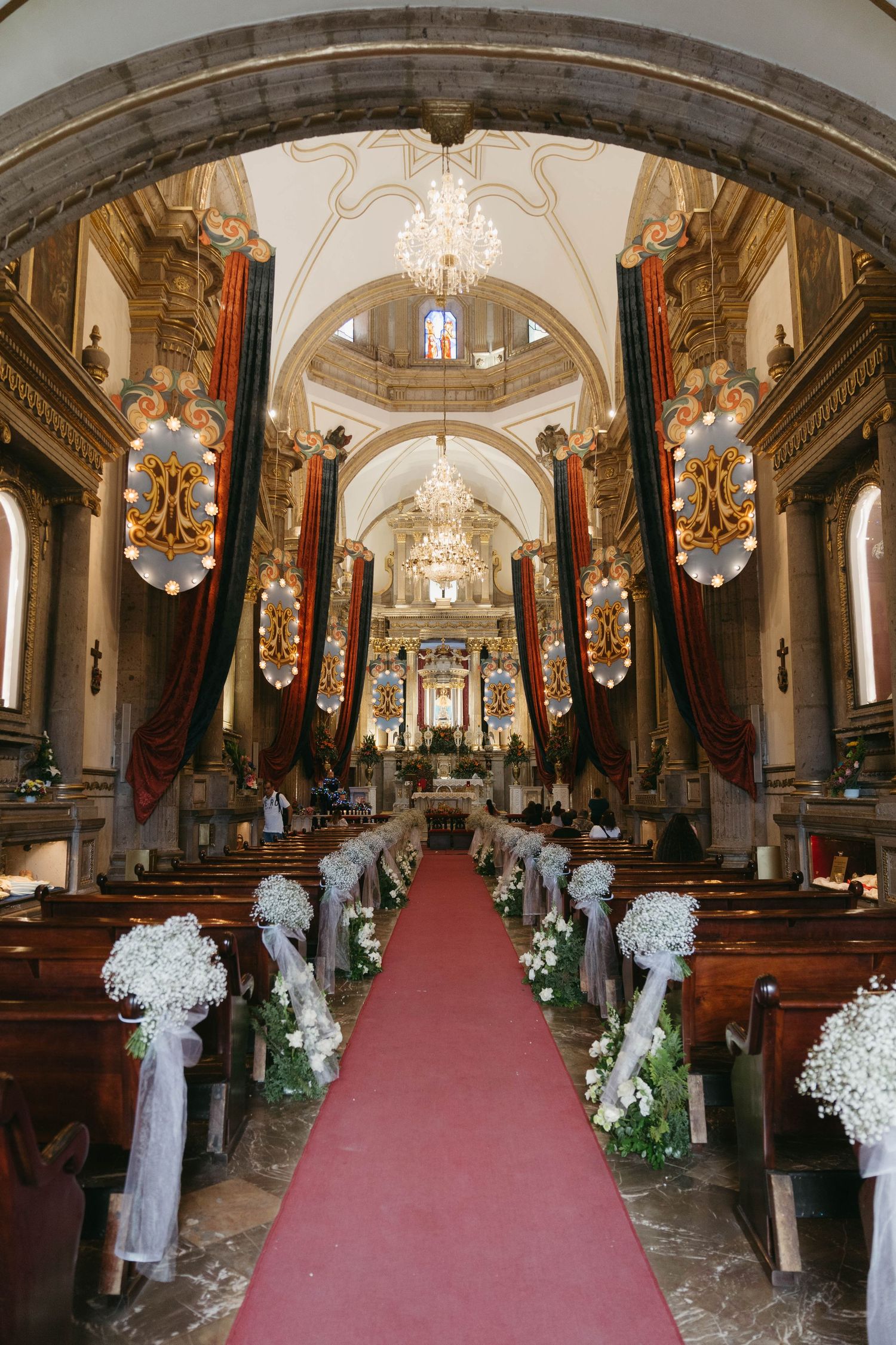 Ornate church interior decorated with white floral arrangements and red carpet for a wedding ceremony.