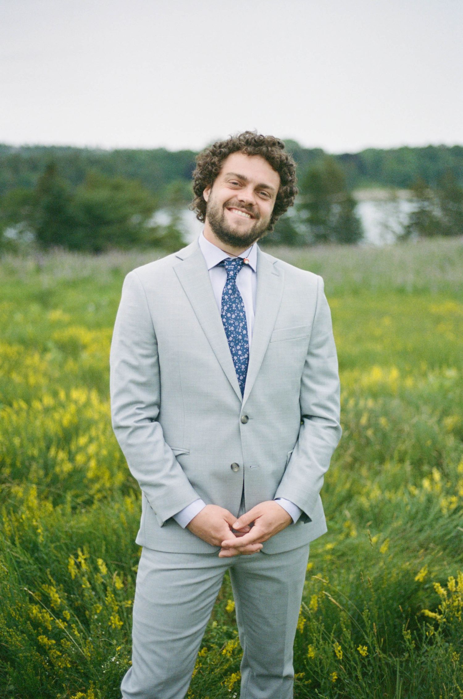 Man in light gray suit with blue tie stands in field of yellow wildflowers.