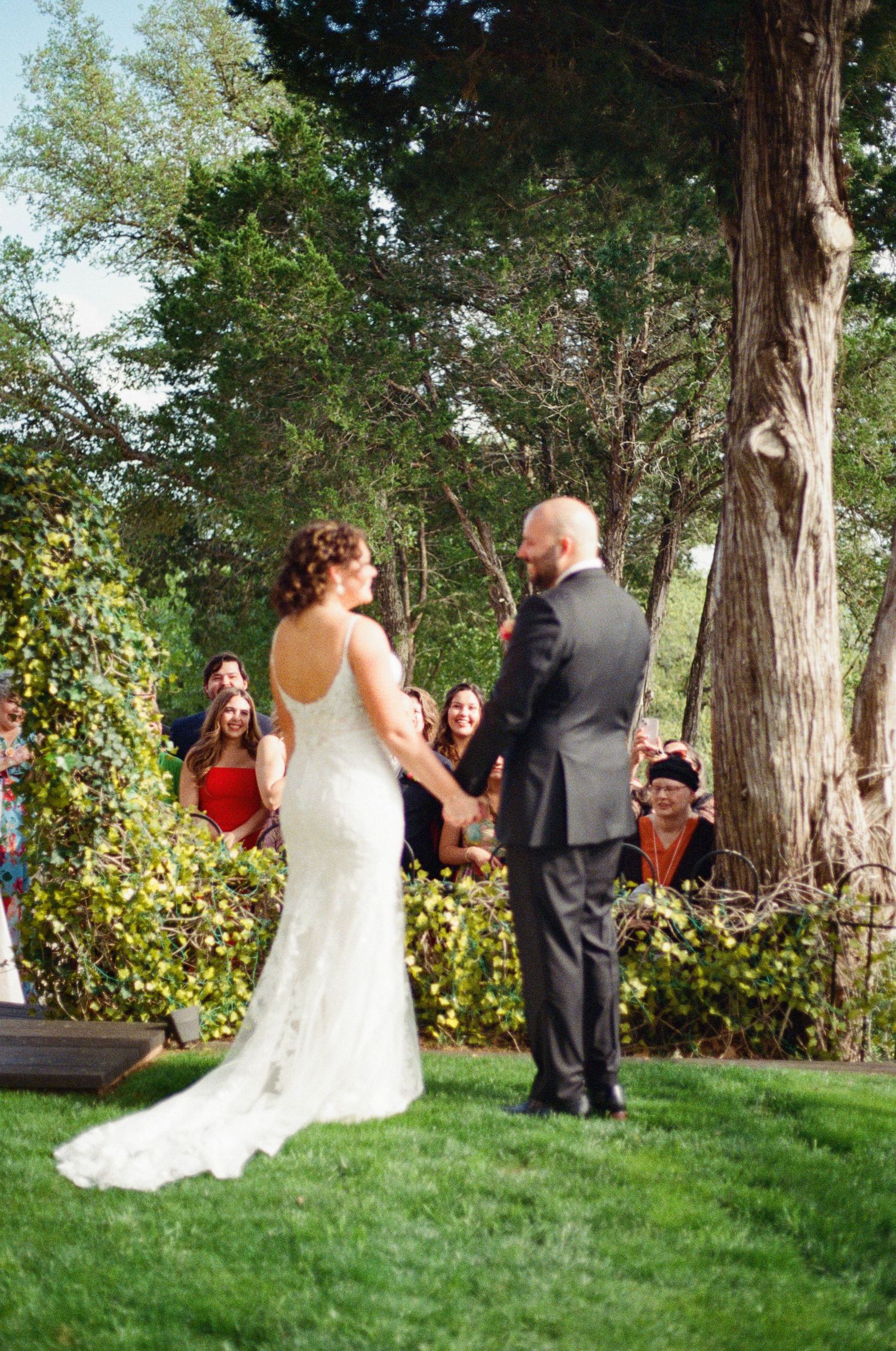 An outdoor wedding ceremony taking place in a lush garden setting with tall trees in the background.