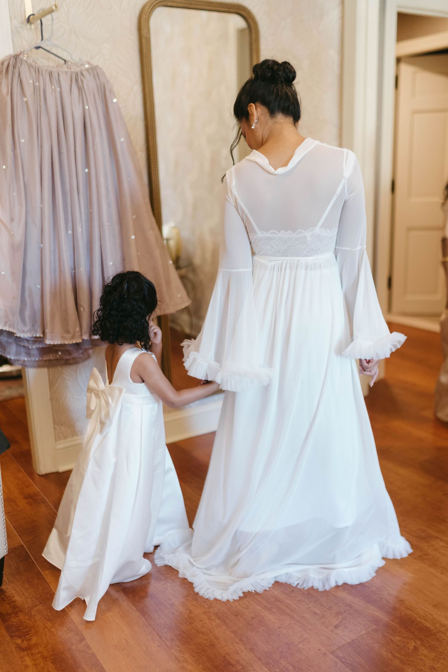 Bride in white gown holding hands with flower girl in matching white dress with bow, viewed from behind in elegant room.