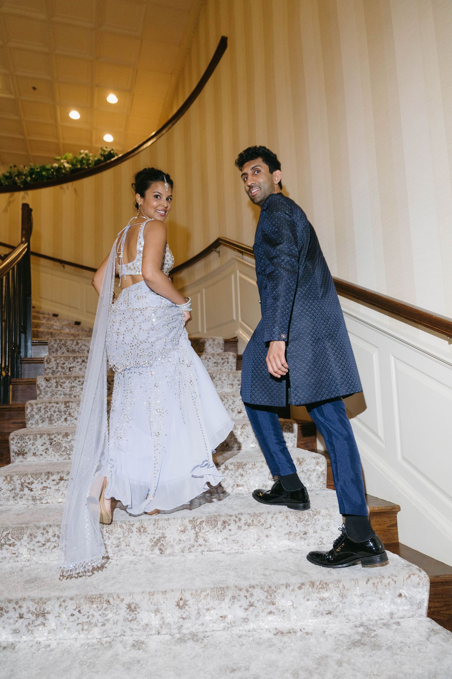 Indian couple in traditional wedding attire posing on elegant staircase, bride in silver lehenga and groom in blue sherwani.