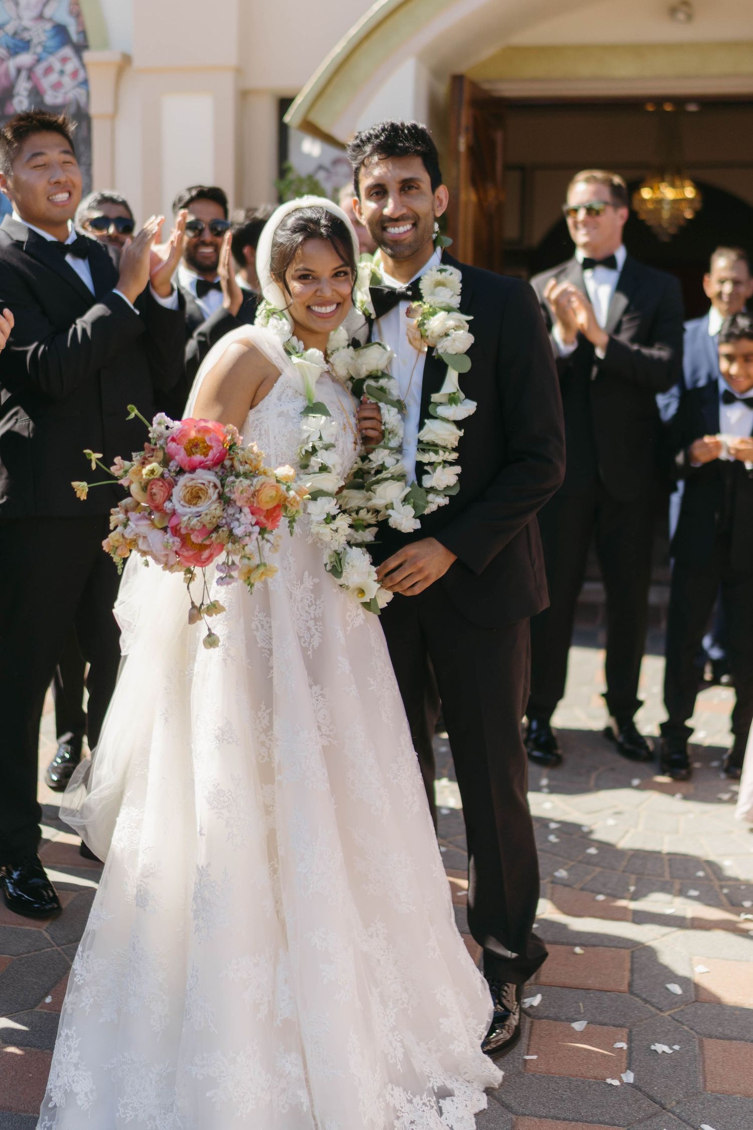 A smiling bride and groom exit a church, with the groom wearing a floral lei and the bride holding a vibrant bouquet.