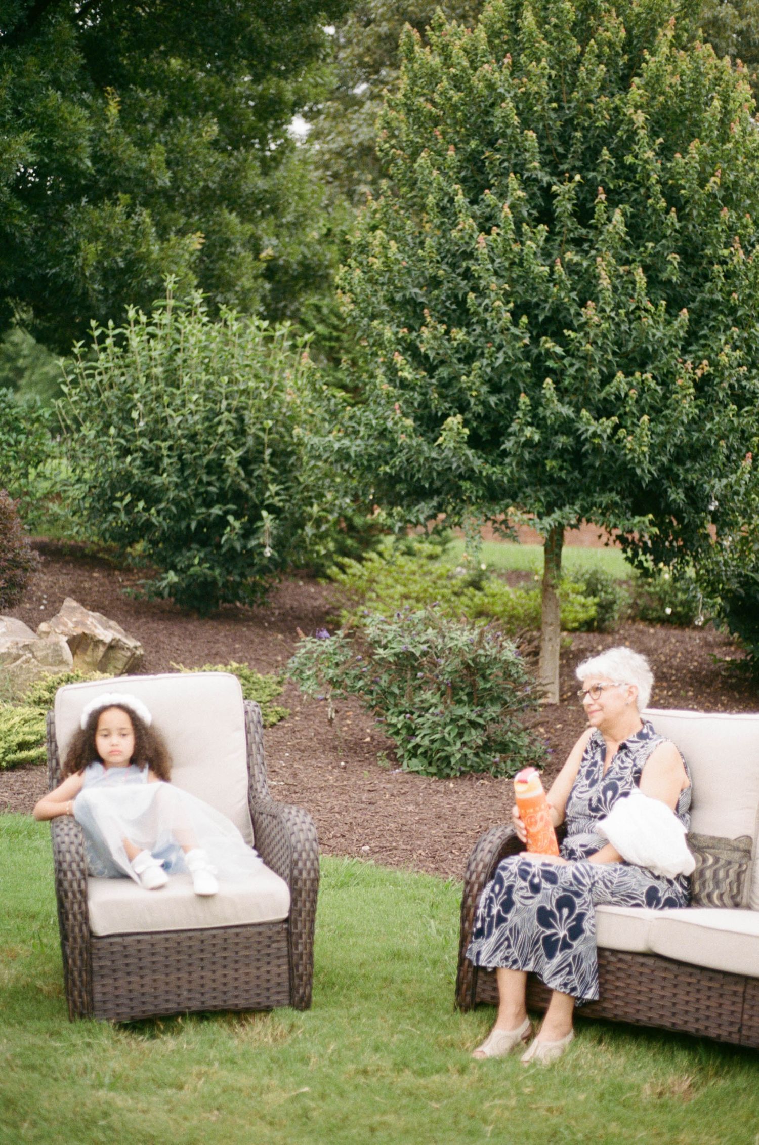 Young girl and elderly woman relax on wicker outdoor furniture surrounded by lush green garden landscaping.