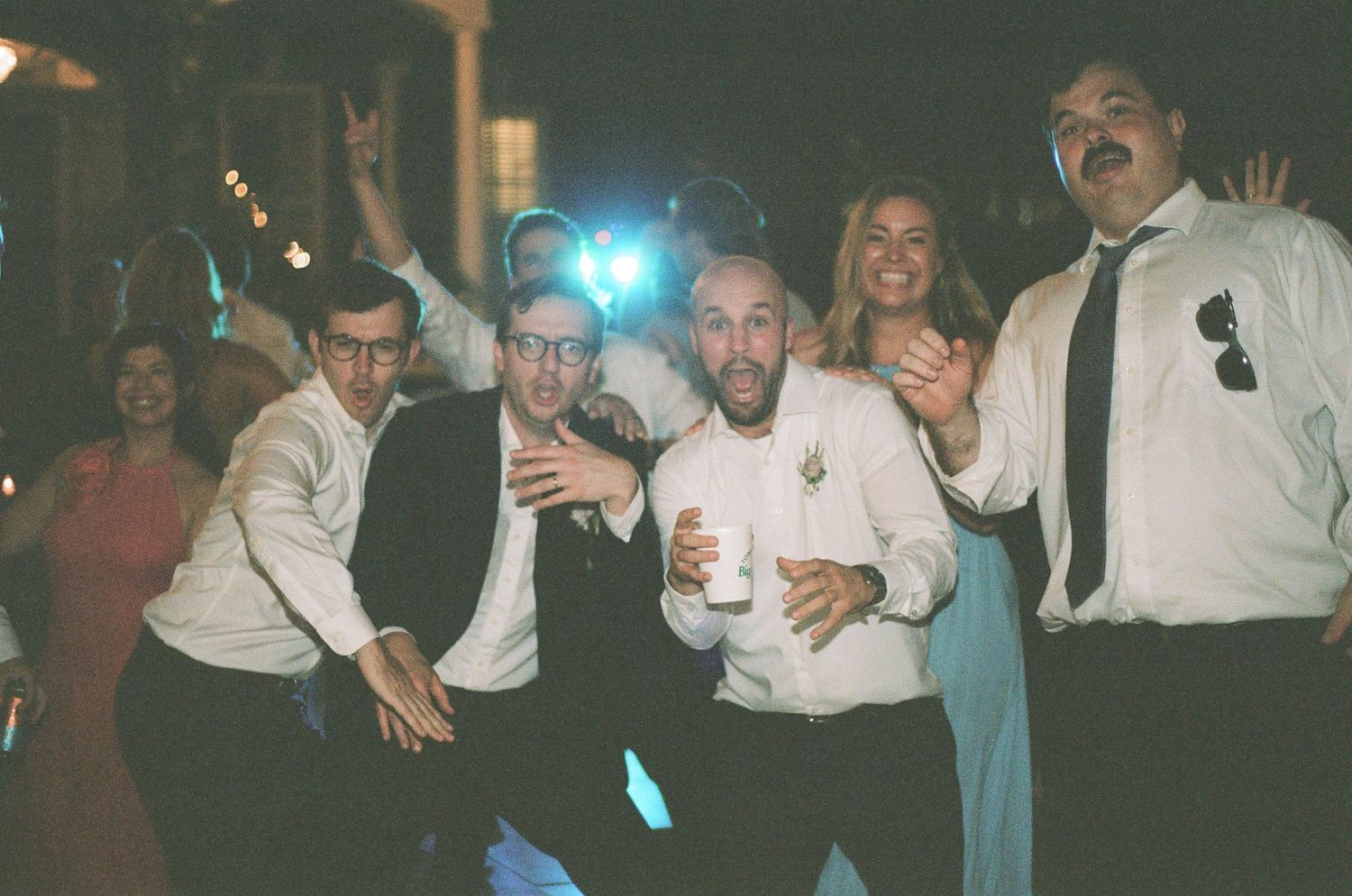 Groomsmen and wedding guests celebrate energetically on the dance floor at a lively wedding reception.