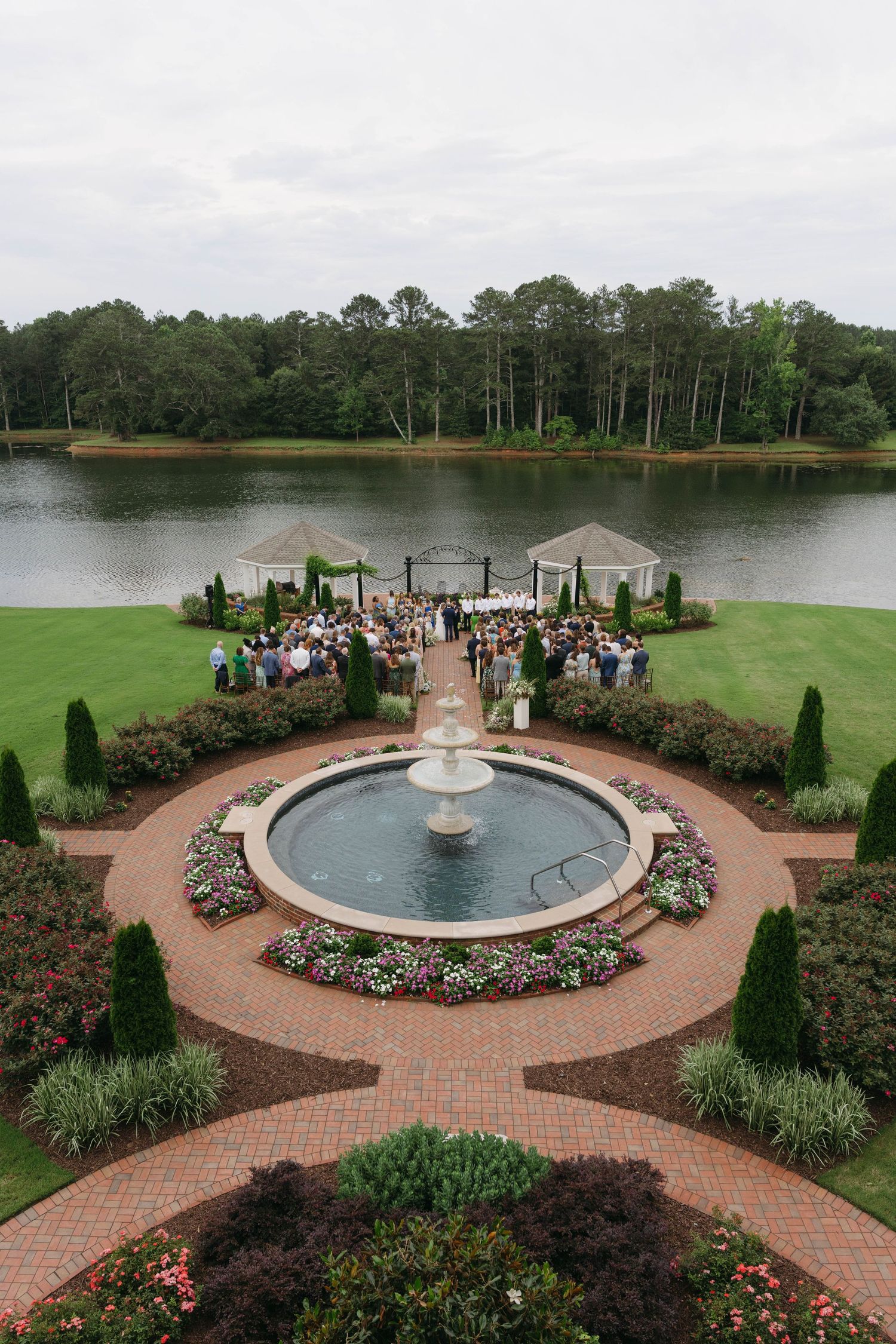 Aerial view of outdoor wedding ceremony by a lake with fountain and manicured gardens.