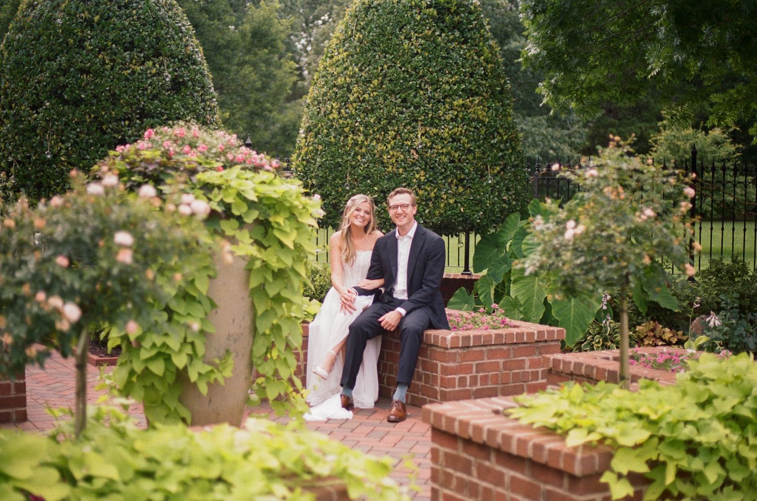 Smiling bride and groom pose together in lush formal garden with manicured hedges and brick walls.