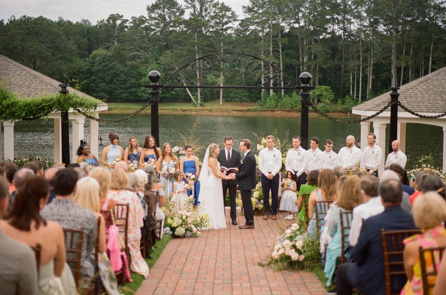 Outdoor wedding ceremony on brick patio with bride and groom at altar, guests seated, lake and trees in background.