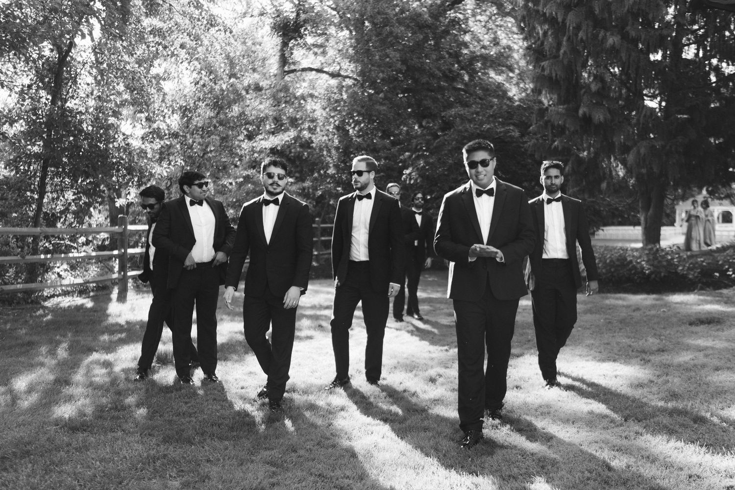 A groom and groomsmen in black tuxedos and bow ties walk together outdoors in a black and white photo.