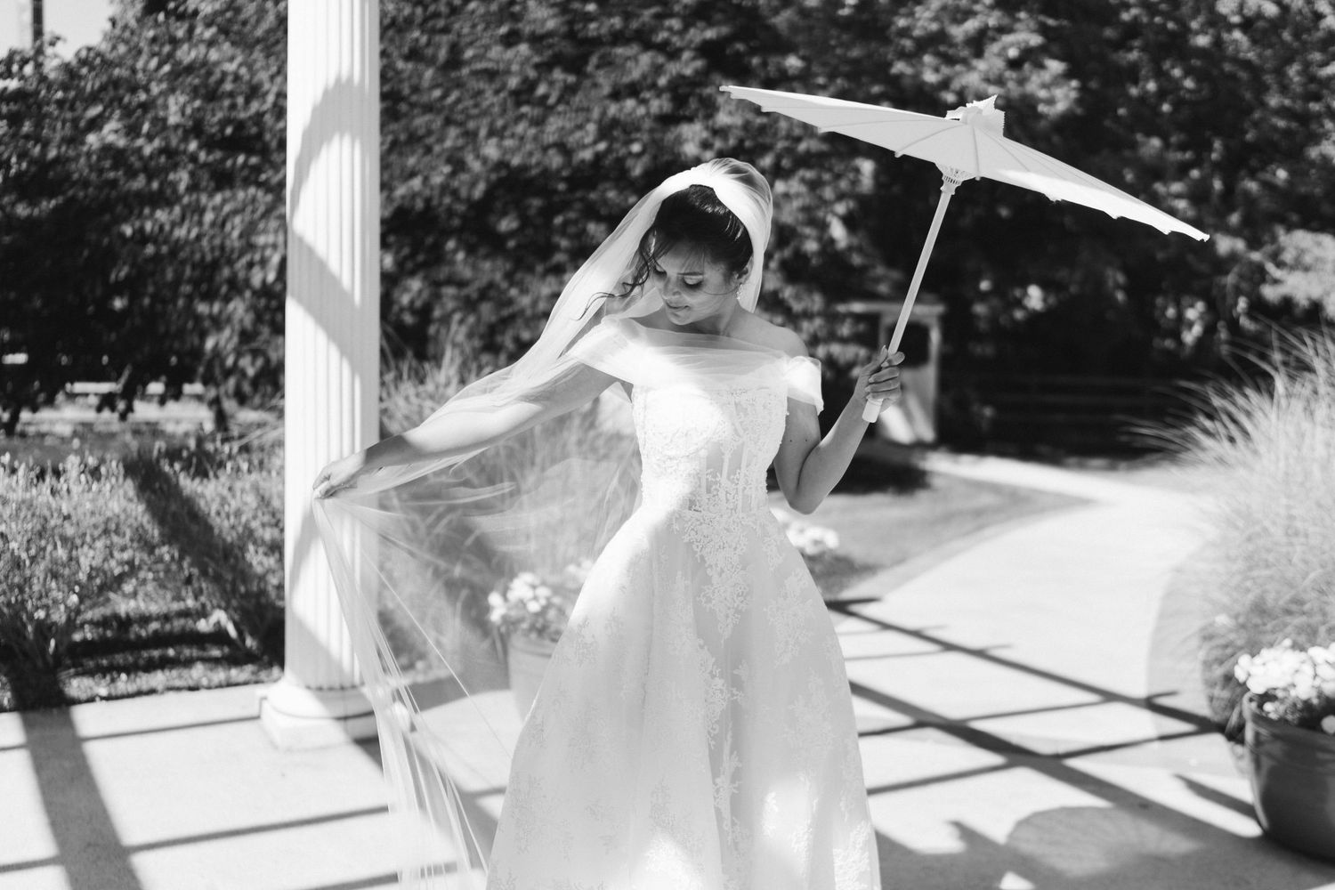 Black and white photo of a bride in a white gown holding a parasol and bouquet, veil flowing in the breeze outdoors.